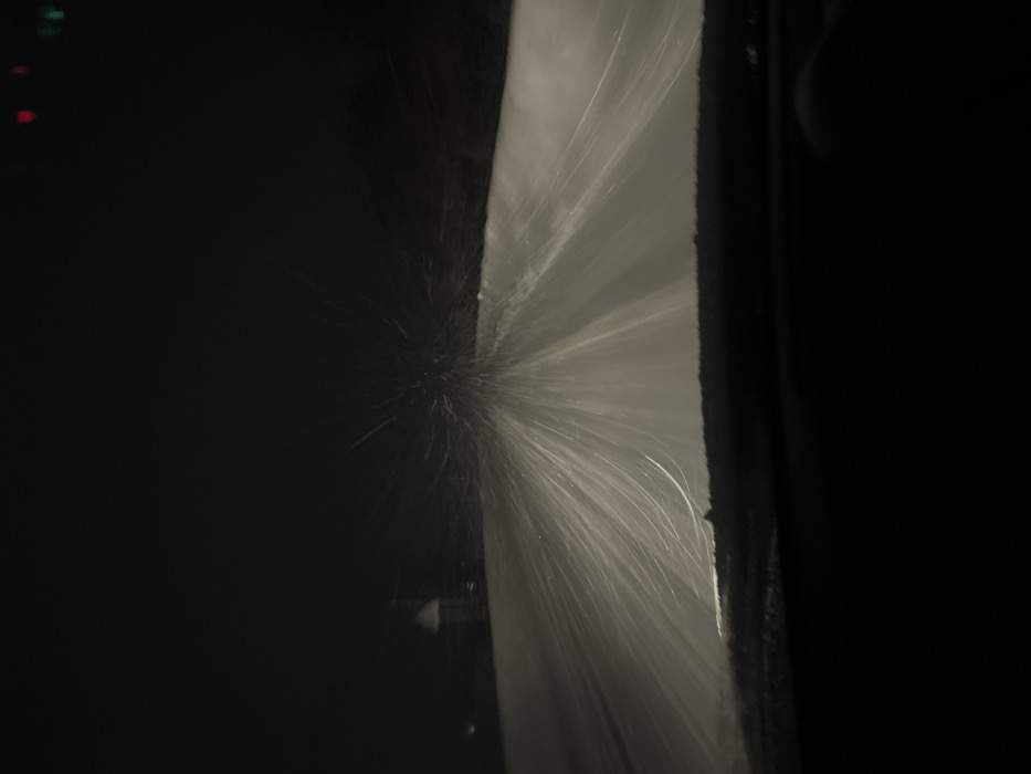 This image captures a nighttime scene in Davydkovo, Russia, viewed from inside a vehicle during a heavy snowfall. The lower foreground shows the dark top of a vehicle dashboard. Intense, illuminated snowfall forms numerous streaks of light across the midground, reflecting light from the vehicle's headlights and extending forward onto a snow-covered road. Visibility is significantly reduced by the falling snow. In the distant background, to the left, a dimly lit building structure with a pointed roof or spire is discernible, partially obscured by snow. Faint red and green lights are visible in the far upper right corner, suggesting distant traffic signals or other light sources. No individuals are visible. The environment indicates severe winter weather conditions.