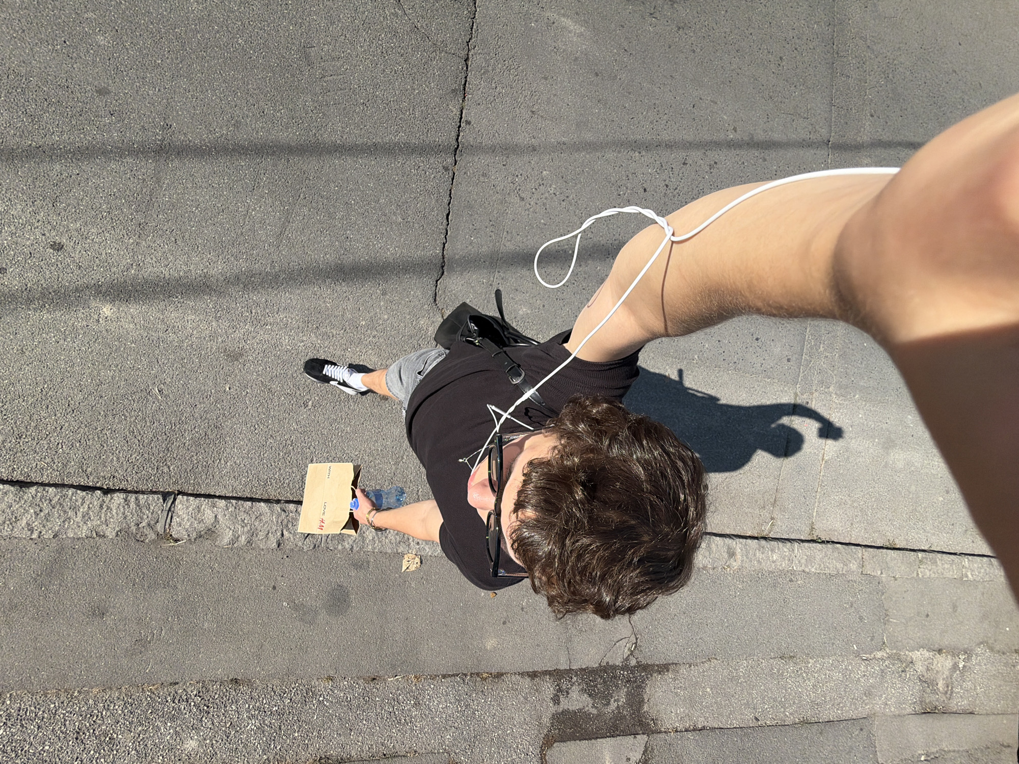 The scene depicts an overhead view of a male pedestrian walking on an urban street surface within the Palilula Urban Municipality, Serbia. The individual, seen from above looking down, has dark, curly hair and wears dark-framed eyeglasses. He is dressed in a black t-shirt, light grey shorts, and black and white sneakers. In his left hand, he carries a small brown paper bag with "LOVE H&M" text visible upside down, containing a blue-capped plastic water bottle. White wired earphones are connected, with the cable looped and tied around his right forearm. A black sling bag or small backpack is partially visible on his left side. The ground consists of a textured, light grey gravel strip on the far left, bordering a raised, rougher stone transition, which then leads to a dark grey asphalt surface. A prominent horizontal crack runs across the asphalt path in the middle distance. Long shadows cast by the person and unseen vertical objects suggest bright overhead sunlight.