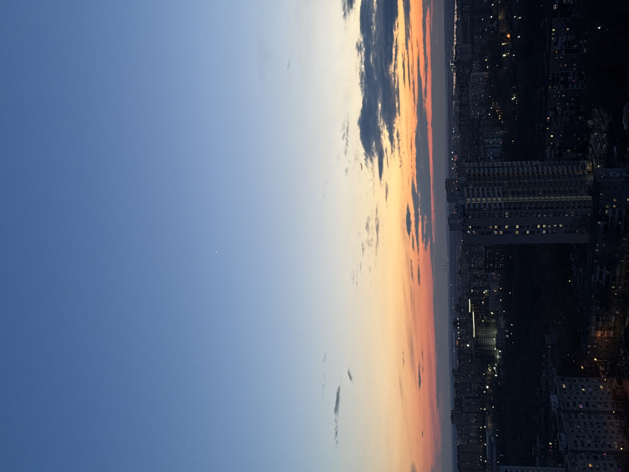 The image presents a wide, expansive view of a city skyline at twilight, observed from a high vantage point. The upper two-thirds of the frame are dominated by the sky, displaying a beautiful gradient from a clear, deep blue on the left to warm oranges and yellows along the horizon line on the right, suggesting either sunset or sunrise. A band of dark, scattered clouds is visible across the center-right portion of the sky, contrasting with the luminous colors. A solitary, faint white dot, likely a star or planet, is noticeable in the upper left quadrant of the blue sky.
The lower third of the image depicts a sprawling urban landscape. Numerous buildings, including what appear to be high-rise residential or commercial structures, form a dense mass of dark silhouettes against the brighter sky. Many windows are illuminated, and streetlights or other urban lights create a myriad of small, bright points scattered across the cityscape, hinting at the start of evening activity. No people or specific events are discernible. The scene evokes a sense of calm and vastness. There is no visible text in the image. The location context indicates this is a view of Perm, Russia.