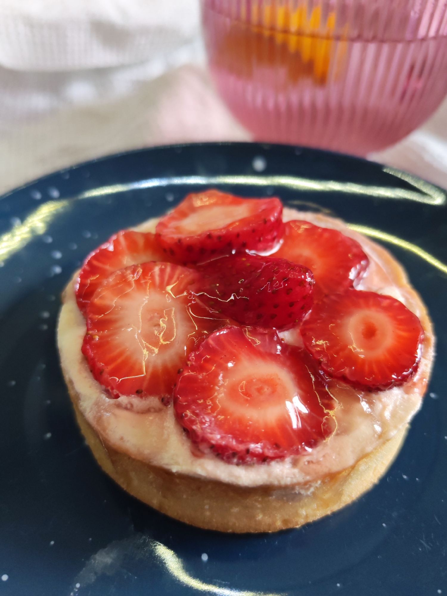 A close-up photograph captures a strawberry tart on a dark blue plate. The tart features a golden crust, a creamy filling, and sliced strawberries covered in a glaze. A pink glass filled with a clear liquid, possibly a drink, stands in the background to the upper right. A white or light-colored cloth provides a blurred background behind the plate. The location and specific details about Izhevsk, Russia, are not discernible from the provided image.