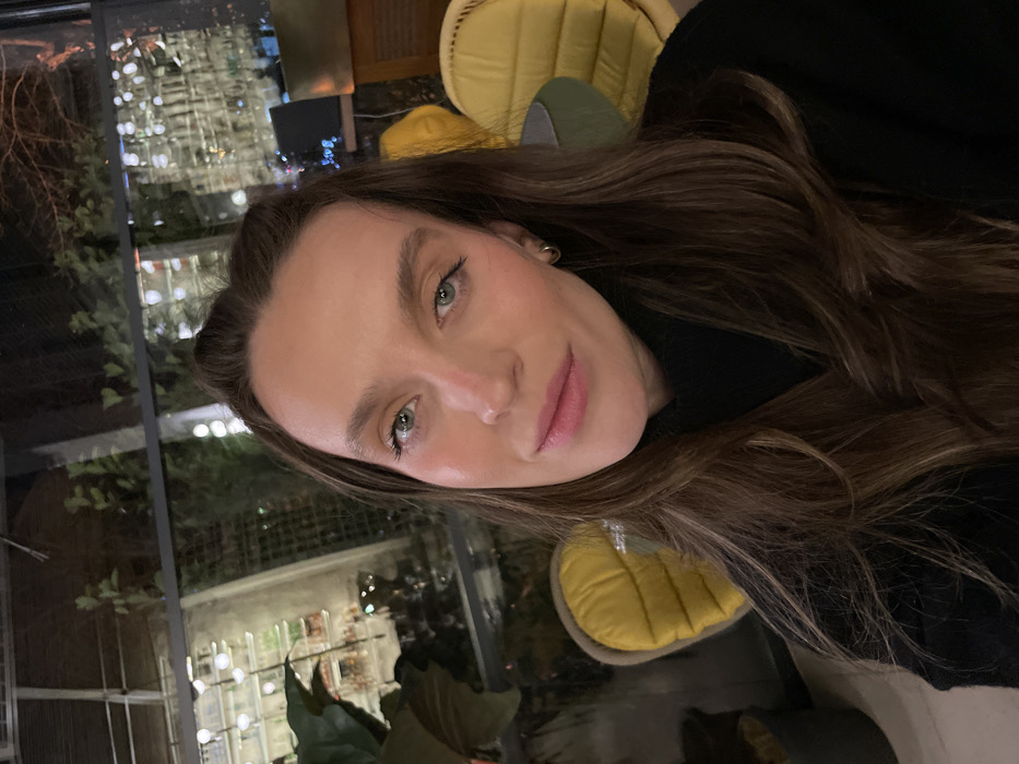 A woman with long, brown hair and light green eyes poses for a selfie. She has fair skin and pink lips. She wears a black turtleneck sweater and gold hoop earrings. The background appears to be a restaurant with yellow chairs and outdoor lighting visible through a window. The photo has a slightly tilted perspective.