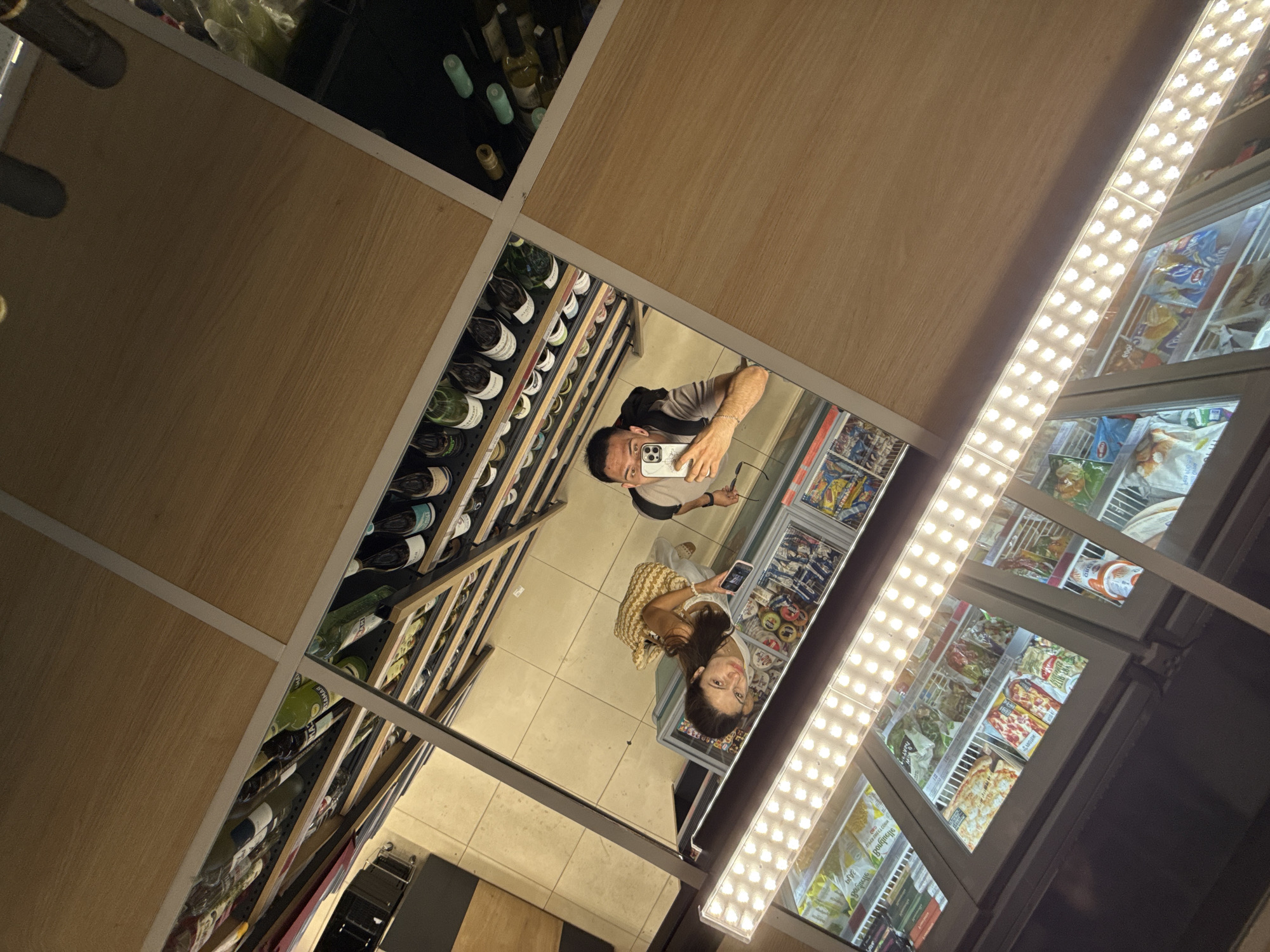 A reflection in a mirror shows a young couple taking a selfie in what appears to be a well-lit grocery store or convenience store. The man is holding a smartphone, wearing a grey t-shirt and a backpack, and looking at the camera. The woman is standing beside him, wearing a white top and holding a straw bag and a pink smartphone. They are positioned in front of a refrigerated display filled with frozen food items, including pizzas and bagged vegetables. Above them, shelves stocked with bottles of wine and other beverages are visible. The store features wooden paneling and bright LED lighting. The scene is consistent with a commercial establishment in Varna, Bulgaria, potentially a supermarket or a specialized food store.