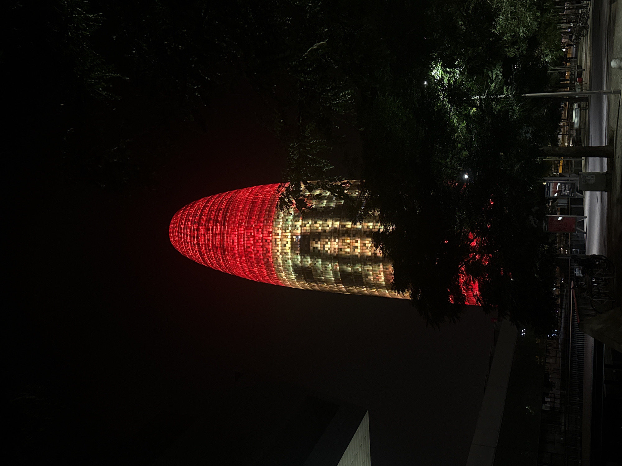 The image captures the iconic Torre Glòries skyscraper in Barcelona, Spain, at night. The building is illuminated with red and golden lights, creating a striking visual against the dark sky. The lower portion of the tower displays a pattern of golden lights, while the upper section is dominated by vibrant red illumination.
In the foreground, several trees with dense foliage partially obscure the view of the tower and the surrounding area. The ground is paved, and in the middle ground, there's a pathway with streetlights and some parked bicycles. A dark, modern building with a slanted roof is visible to the left of the tower, partially in shadow. The overall scene conveys a nighttime urban environment with the illuminated skyscraper as the central focal point. There are no visible people in the frame.