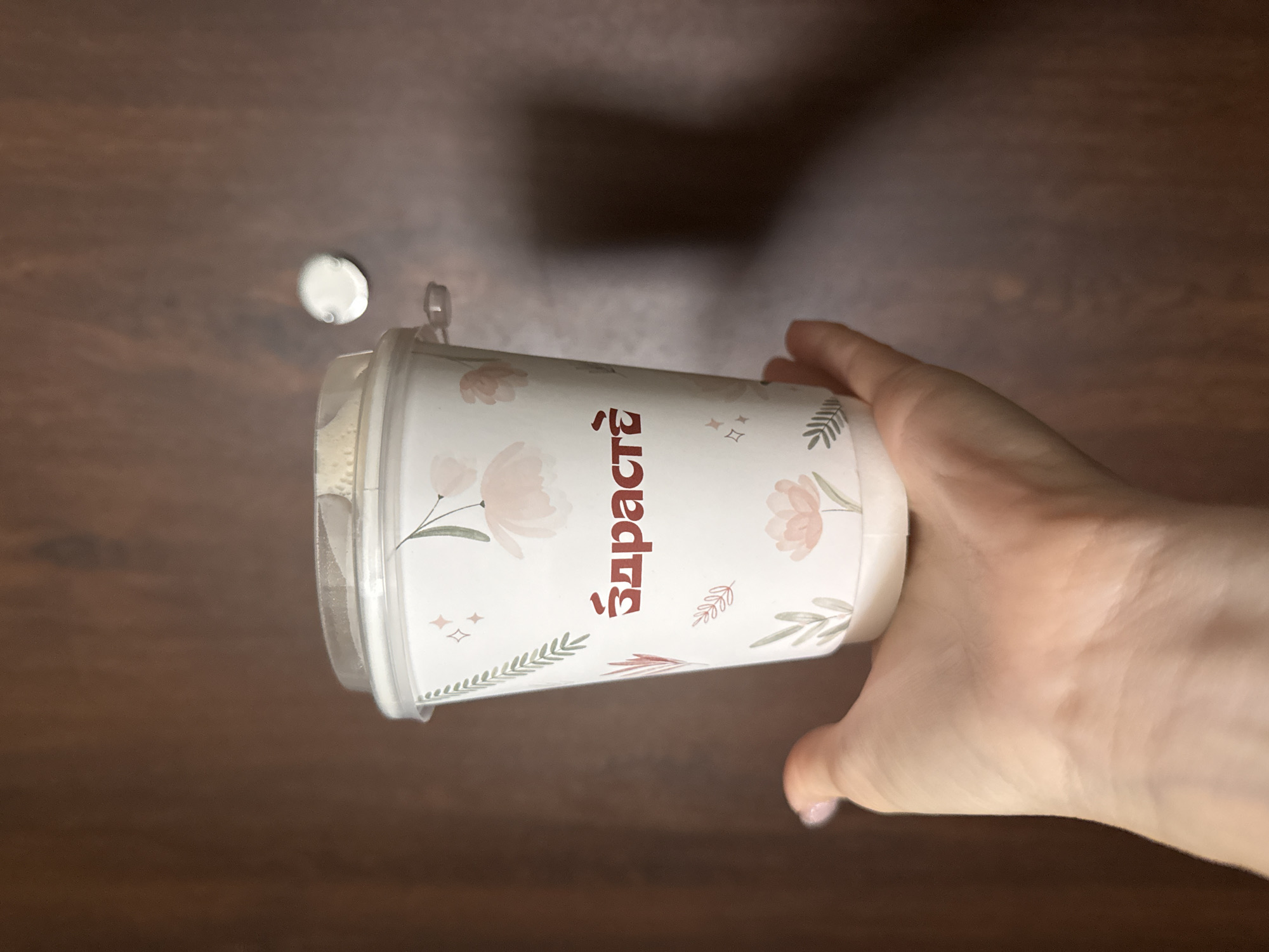 A hand holds a paper coffee cup adorned with a floral pattern and the word "Здрасте" in red lettering. The cup has a clear plastic lid. The background is a wooden surface with a faint reflection of light. The scene is indoors, with no discernible Moscow landmarks or urban environment visible.