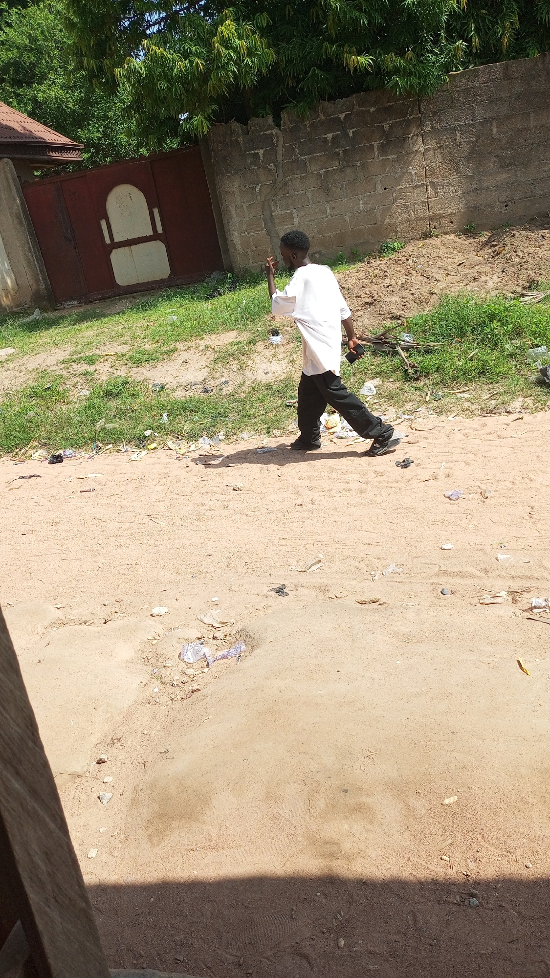 A young Black male is walking away from the camera on a dirt path. He is wearing a white long-sleeved shirt and black pants, and he is holding a dark-colored mobile phone in his right hand. The path is lined with dry grass and scattered debris, including plastic bags and wrappers. In the background, a large, weathered stone wall separates the path from a residential area. A tall, reddish-brown metal gate is visible to the left of the wall, partially obscured by a building with a brown roof. Lush green trees are visible above the wall and gate. The scene appears to be outdoors in a somewhat neglected urban environment. The lighting suggests it is daytime. This location is identified as Jalingo, Nigeria.
