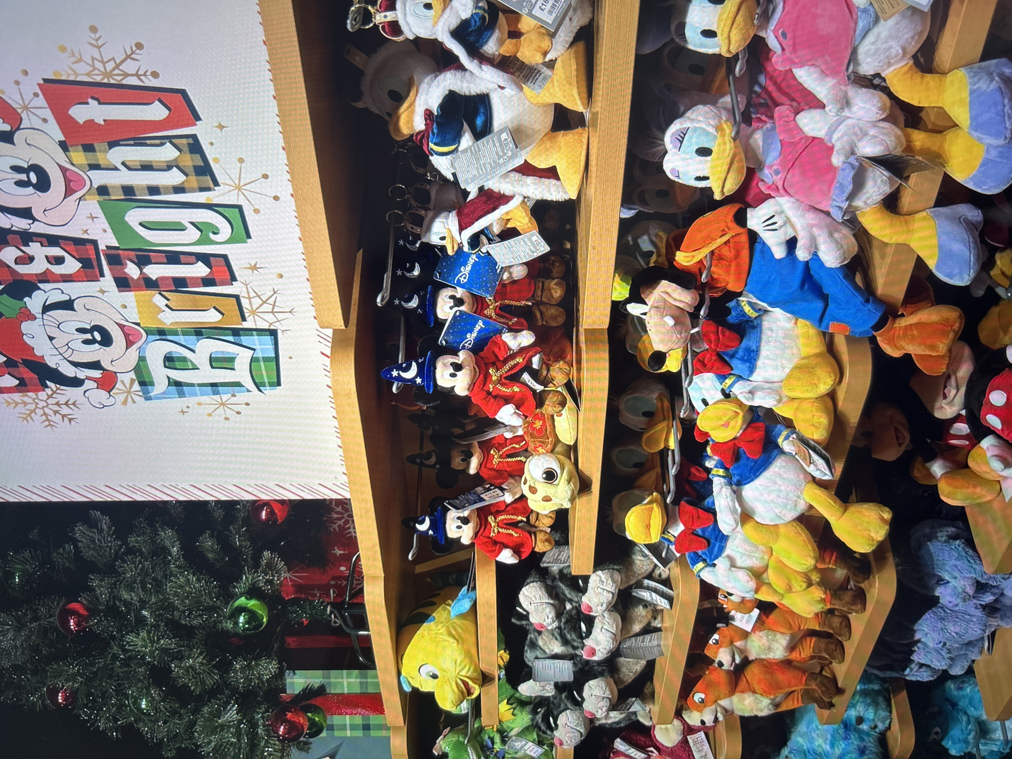 The image shows a display shelf filled with Disney character plush toys. The toys include Mickey Mouse, Donald Duck, and others. A Christmas tree is visible in the lower left corner of the image. A sign with "Bright" on it is next to the shelves with the toys. The image was taken indoors, most likely in a retail store. There is no evidence to suggest that the photo was taken in Gorna Oryahovitsa, Bulgaria.