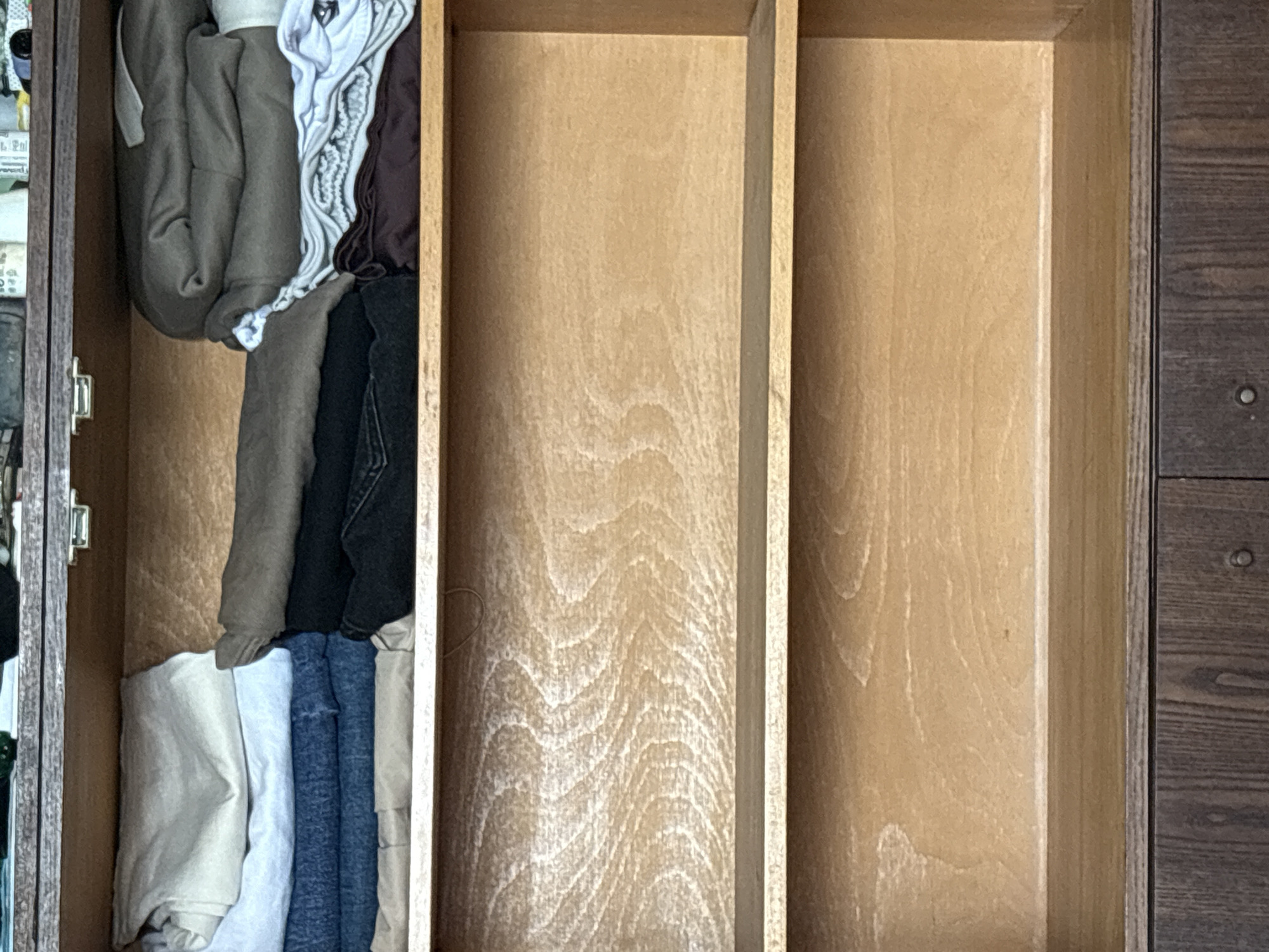 A top-down view shows a partially filled wooden drawer with two empty compartments. The left compartment contains folded clothing items stacked on top of each other. The middle and right compartments are empty. The exterior of the drawer and cabinet are also made of wood. The scene location is unknown.