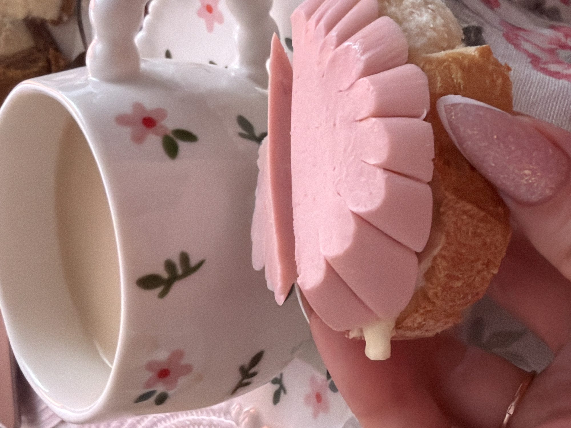 A close-up shot shows a partially consumed sandwich held in a person's hand, positioned adjacent to a white teacup. The sandwich consists of slices of pink sausage arranged in a radial pattern on a bread roll, with a visible dollop of white sauce. The teacup is adorned with a floral pattern featuring pink flowers and green leaves. The person holding the sandwich has a manicured fingernail with a white tip. The background shows a floral tablecloth or bedding. While there is no definitive information to confirm the location in Saint Petersburg, Russia, the image predominantly features common objects and foods that are not exclusive to a particular region. It is not possible to confirm the location of the image to Saint Petersburg, Russia.