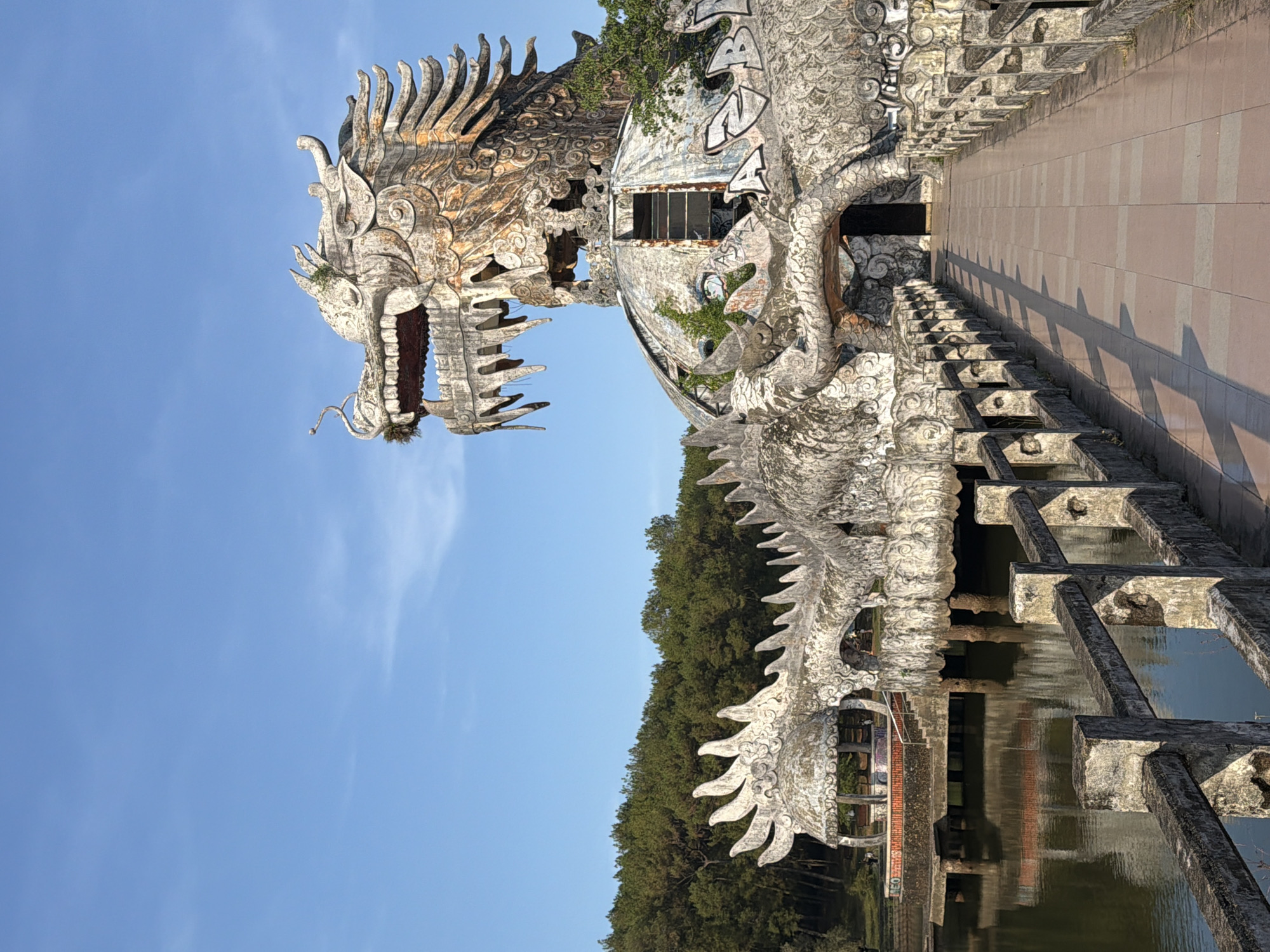 The image depicts a large, weathered, white dragon sculpture in Thủy Xuân Ward, Vietnam. The dragon's head is prominently displayed against a clear blue sky, with the rest of its body extending along a bridge or walkway over a body of water. The sculpture appears to be made of concrete or a similar material, showing signs of age and decay, including discoloration and graffiti. The bridge's structure is visible beneath the dragon's body, with supporting pillars in the water. Lush green trees fill the background, suggesting a natural setting.