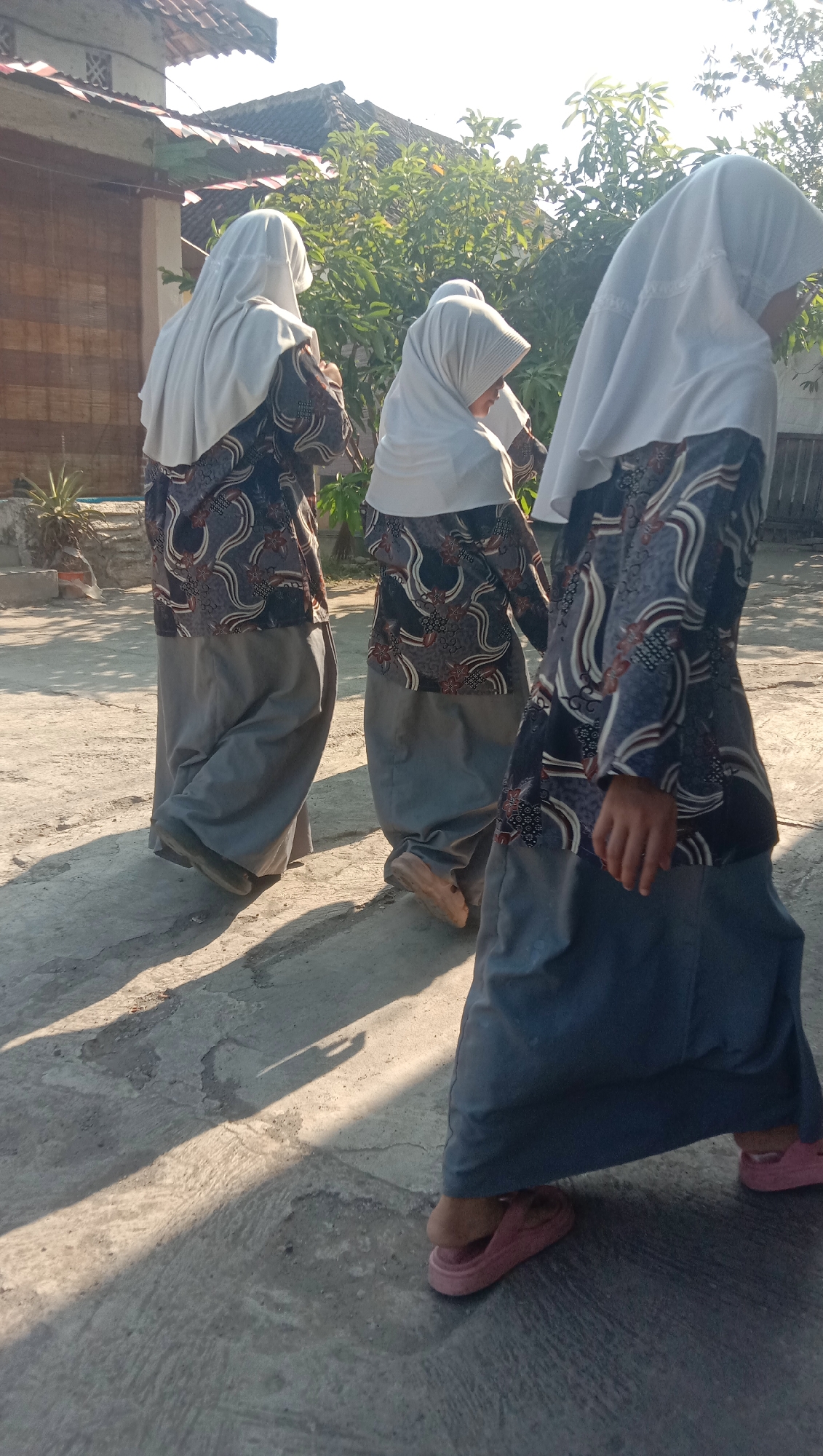 Three individuals, likely students, are walking outdoors. They are wearing light-colored hijabs and patterned tops over long, gray skirts. The ground is a concrete surface with visible cracks and patches. Sunlight casts long shadows from the people and nearby trees. In the background, there is a building with a tiled roof and some vegetation. The scene appears to be in Sidogiri, Indonesia, possibly near a school or residential area.