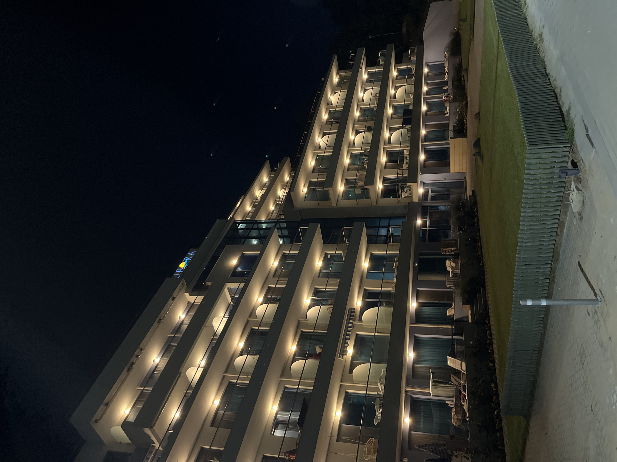 The image shows a multi-story hotel at night, located in Obzor, Bulgaria. The hotel has multiple balconies with railings, and many windows are illuminated from within. The hotel signage, "AURORA," is visible at the top of the building, lit in blue and yellow. A well-maintained lawn is in front of the hotel, bordered by a wooden fence. A paved walkway runs in the foreground, and a gray metal pole stands on the walkway. The overall scene depicts a nighttime view of a hotel establishment.