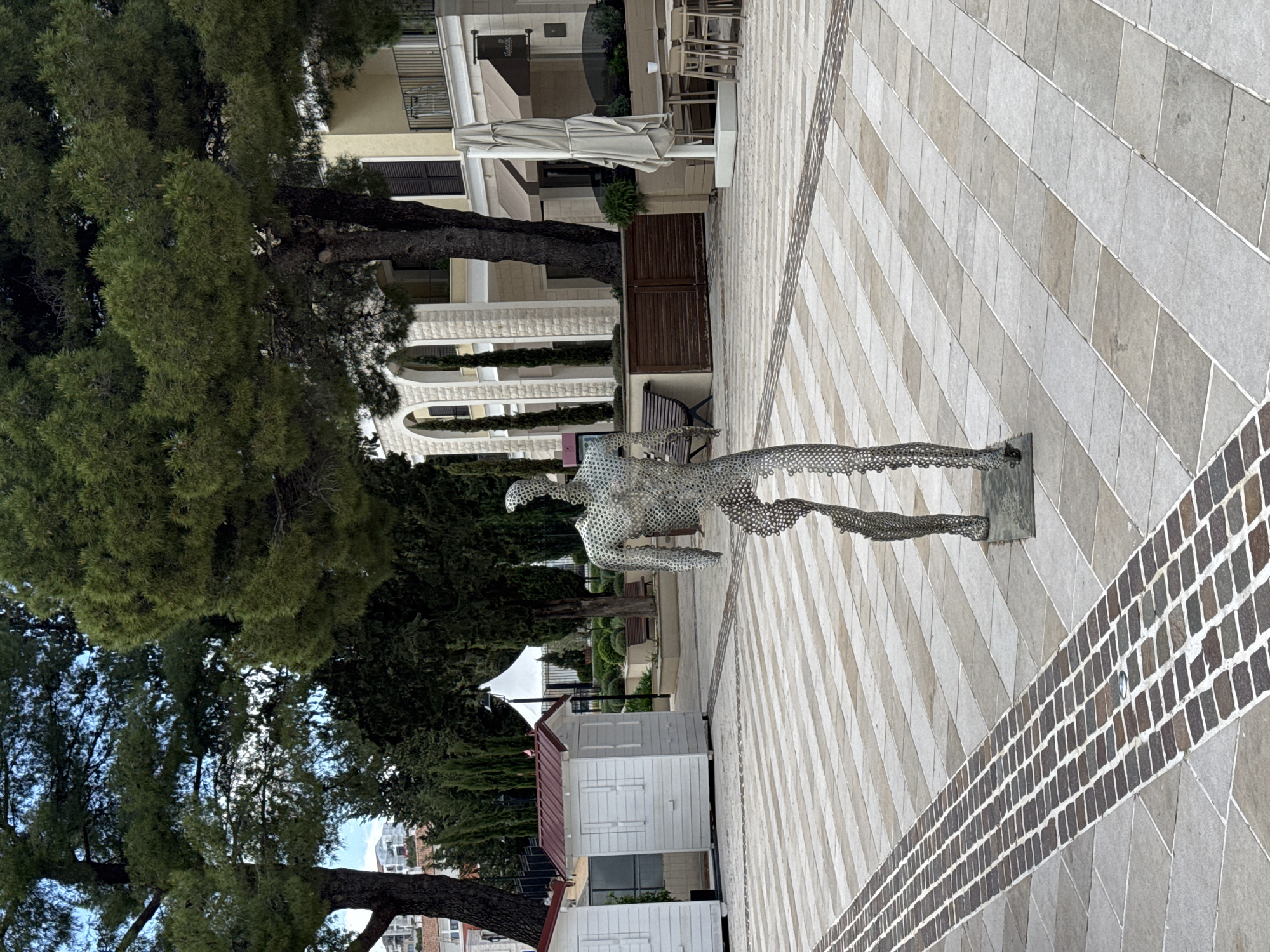 The image displays an outdoor scene in Radovići, Montenegro, centered around a striking metallic sculpture of a human figure in motion, possibly running or lunging. The sculpture is crafted from numerous interconnected metal pieces, creating an open, textured form. It stands on a geometrically patterned paved ground, which features alternating diagonal stripes of light and slightly darker grey or beige rectangular tiles. A distinct diagonal band of darker brown, brick-like paving also traverses the ground.
Tall, lush green trees, likely pines or similar conifers, frame the scene, their dense foliage forming a natural backdrop on the left and extending into the background. Through gaps in the canopy, a clear sky is visible. Behind the trees and towards the right, several light-colored buildings with Mediterranean architectural characteristics are visible, including arched openings, balconies, and dark brown window shutters.
Additional elements include a closed, light-colored patio umbrella, a dark wooden bench, and a brown wooden gate or partition. Further to the left, two small, white, hut-like structures with reddish roofs are present, resembling kiosks or small utility buildings. The scene is illuminated by soft daylight, suggesting either an overcast day or indirect light, as no harsh shadows are apparent. There is no visible human activity or discernible text in the image. The overall impression is one of a tranquil, well-maintained public or resort area.