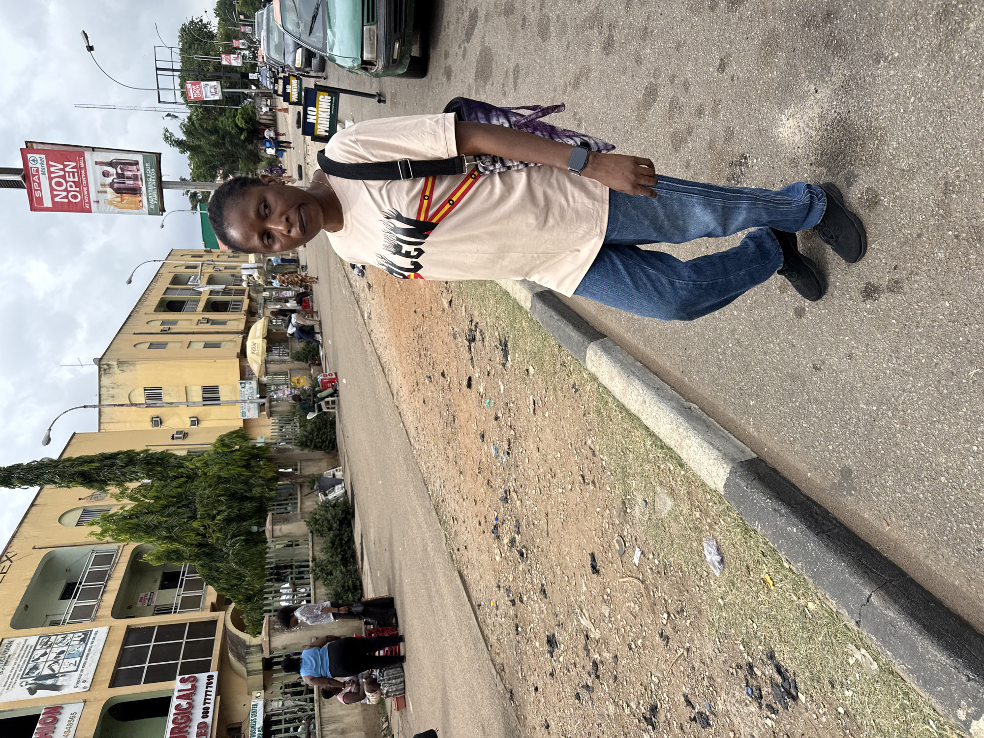 The image shows a woman standing on a sidewalk next to a street in Abuja, Nigeria. She is wearing a light-colored t-shirt with a graphic design, blue jeans, and black shoes. She has a bag slung over her shoulder. In the background, there is a multi-story yellow building with businesses including "SURGICALS" and "VISION CENTER" advertised on its facade. Across the street, there is a large SPAR supermarket sign that reads "NOW OPEN at Novare Central Mall." Other vehicles are visible on the road. The street appears to be busy with pedestrians. There are signs indicating "NO PARKING". The environment is urban with trees and utility poles present.