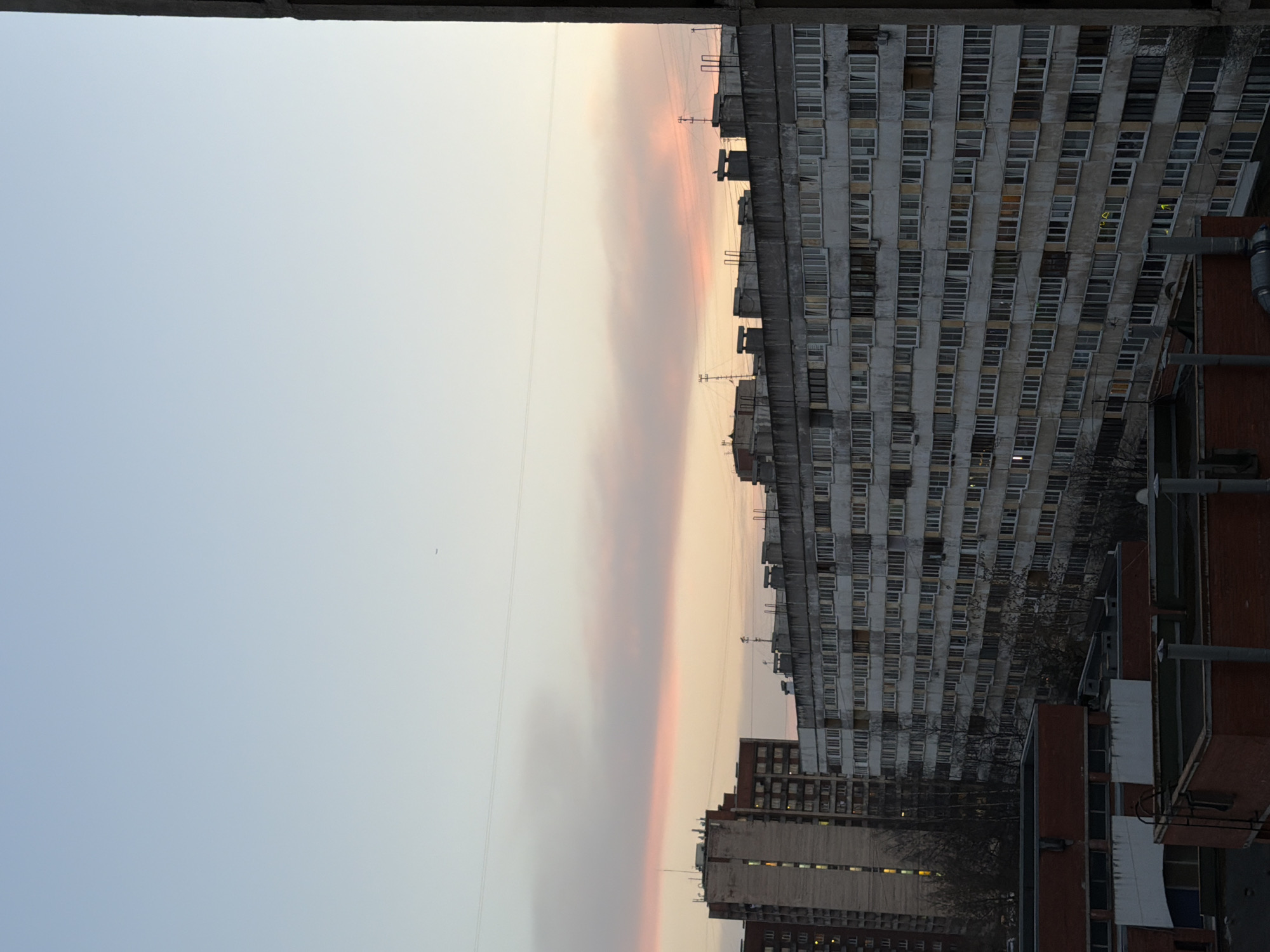 The image shows a view from a high vantage point, likely a building or elevated structure, in what appears to be a densely populated urban area, likely in Russia.
The horizon displays a pale, fading light, indicative of dawn or dusk.
The sky transitions from a soft, gradient gray/pink to a clearer blue-gray, potentially indicating the aftermath of a sunset.
The cityscape is dominated by Soviet-era apartment buildings, which are high-rise and rectangular, feature multiple uniform windows, and are made of concrete.
There are various buildings in the shot, the closest one has a reddish tone, and is most likely made of brick, with dark windows.
There is also a tall building, in gray concrete.
Power lines cut across the sky.
The image is rotated counter clockwise, and taken from a window frame.
Based on the architectural style and the color of the buildings, it is possible that the image was taken in Saint Petersburg, Russia.