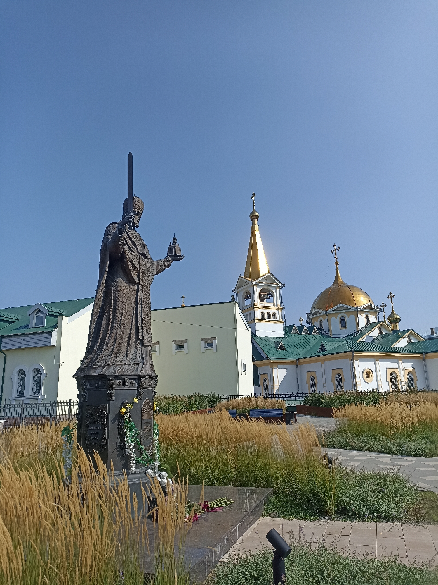 The image captures a view of the Ascension Cathedral and a statue of Saint Nicholas of Myra in Novosibirsk, Russia. The statue, a bronze figure holding a sword and a model of a church, stands in the foreground, surrounded by dry grasses and ornamental plantings. Behind it, the Ascension Cathedral is visible, featuring a prominent golden dome, a bell tower, and a green roof, all set against a clear blue sky. The architecture suggests a historic religious site. The overall scene is peaceful and bathed in daylight.