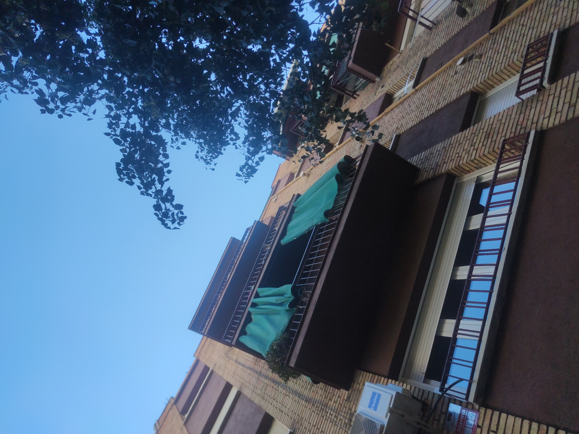 The image depicts the exterior of a brick building with balconies. The building is tan brick with brown trim and dark brown balconies. Some balconies are covered in green material. One balcony is open and some windows are barred. An air conditioner is affixed to the wall on the building. Tree branches with leaves are visible in the upper left corner of the image. The sky is blue and mostly clear. The location appears to be an urban area, city Unknown, Unknown.