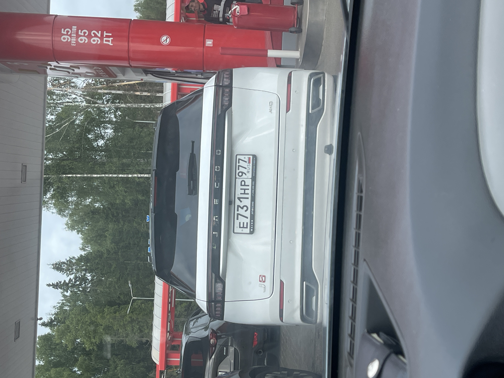 The image shows the rear of a white SUV at a gas station, with a red gas pump to the left and trees and a cloudy sky in the background. The SUV has a license plate that reads "E731HP 977 RUS" and "JAECOO" branding. The gas pump has text indicating fuel types and prices, including "95 EVOLUTION" and "95 92 ДТ". There are also red and white barriers and a person partially visible in the background near the pump. The scene appears to be during daylight, with overcast weather. The overall impression is a common scene at a gas station.