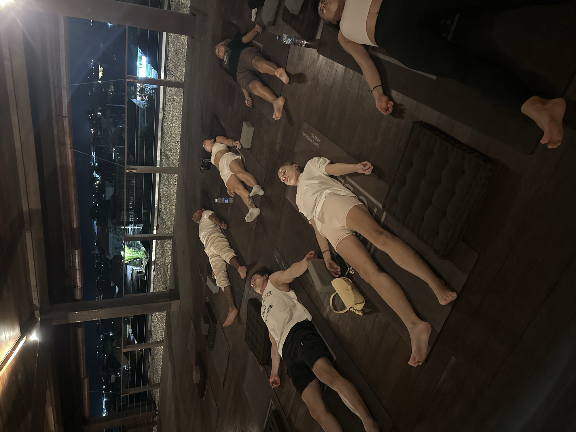 Location: Mengwi, Indonesia. Indoor space with a view of the night cityscape through a window with a metal wire railing.
People: Several individuals lying on yoga mats, seemingly in a relaxation or meditation pose. The individuals are dressed in workout attire.
Objects: Yoga mats, water bottles, yoga blocks, and a small bag.
Actions: People are lying down, possibly meditating or relaxing after a yoga session.
Environment: A room with wooden floors. The room is dark, with only dim lighting, and the cityscape is visible through the window, suggesting it is nighttime.
