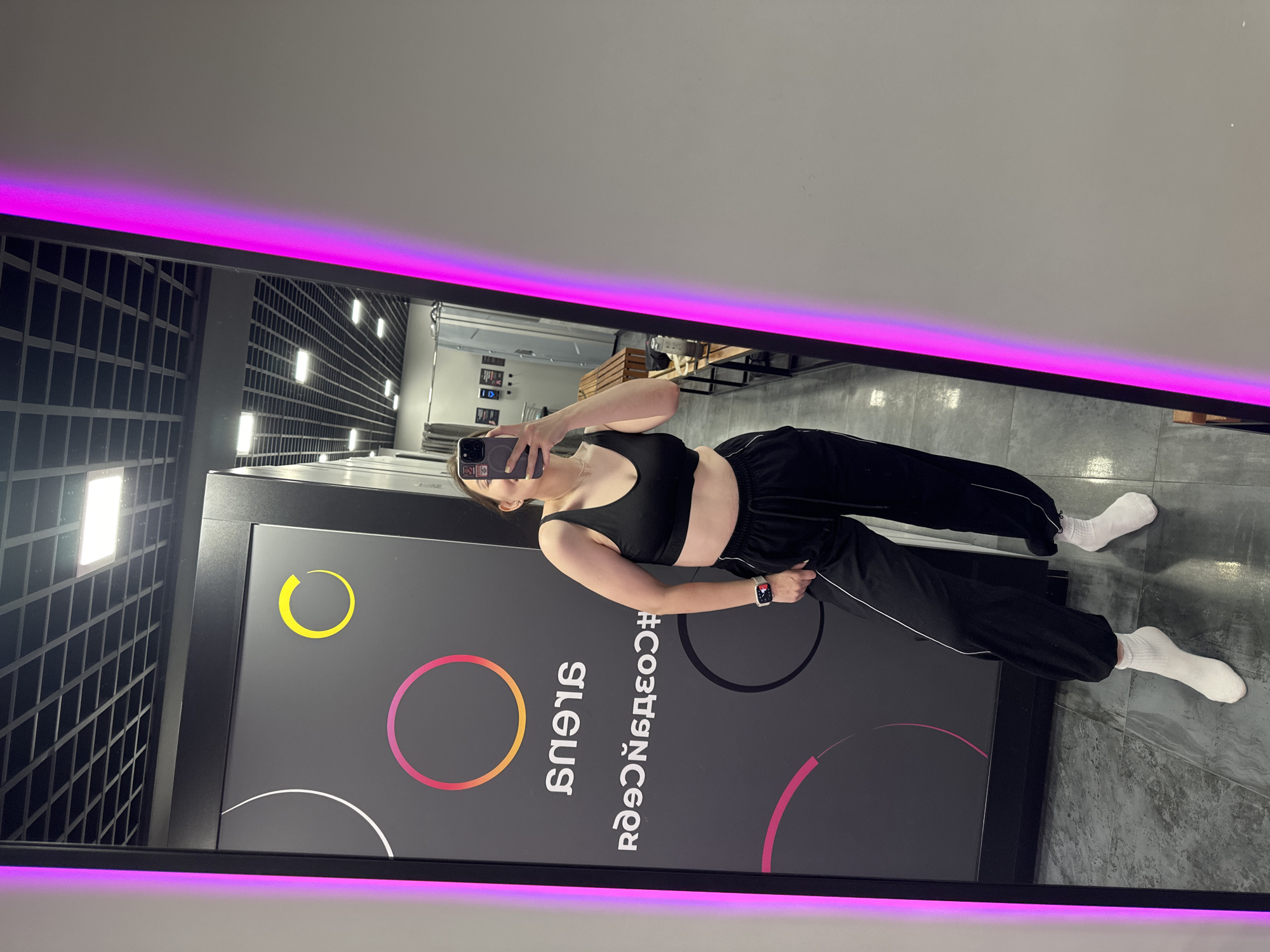 The image is a full-length mirror selfie taken in what appears to be a locker room or dressing area, possibly at a gym or fitness facility. A young woman, dressed in a black sports bra, black sweatpants with white piping, and white socks, is standing and holding a smartphone to take the picture. She is wearing a smartwatch on her left wrist.
The background visible in the mirror shows a dark gray wall with a circular design in white, yellow, and a gradient of pink to orange. There are also some logos or text on the wall, including what appears to be reversed Cyrillic text. Further in the background, a row of wooden benches and what look like shower stalls or changing cubicles can be seen. The ceiling features a grid of lights. The mirror itself has a prominent bright pink neon light strip running vertically along its right edge. The floor is tiled in a gray, slate-like pattern.
The overall impression is a candid, informal snapshot taken in a private or semi-private space. The image does not contain any specific landmarks or visual cues that would definitively place it in Perm, Russia.