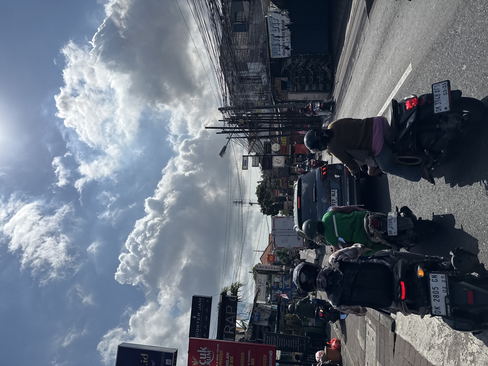 In Denpasar, Indonesia, on a sunny day with dramatic clouds, a busy street scene unfolds. The perspective is from behind a motorcycle, showing a traffic jam with numerous motorcycles and a few cars. On the left side of the street, there are buildings with signs for businesses such as "DOKTER JAM", "TRAP", "PERFUME KSA", and "DENTIST". The right side features more buildings and a dense network of overhead electrical wires. People on motorcycles are wearing helmets, with some carrying passengers. The road surface is asphalt, with white lines marking lanes. The overall atmosphere is one of a vibrant, bustling urban environment.