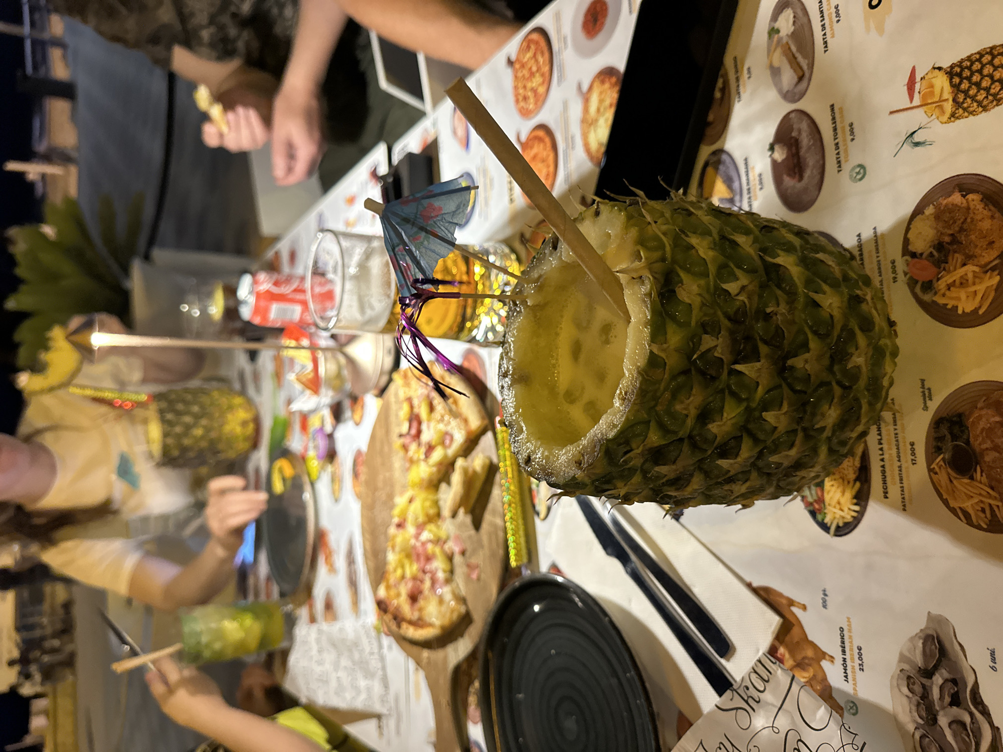 In an outdoor dining setting, a pineapple with a straw and a small umbrella sits on a table. The pineapple contains a yellow drink, and its top has been hollowed out. A wooden pizza peel with partially eaten pizza is visible in the background, along with glasses of drinks, a red can of soda, and cutlery. The table is covered with a patterned tablecloth that appears to be a menu featuring images of food and prices. In the background, out-of-focus figures of people are seated, suggesting a meal is in progress. The lighting indicates it is evening.