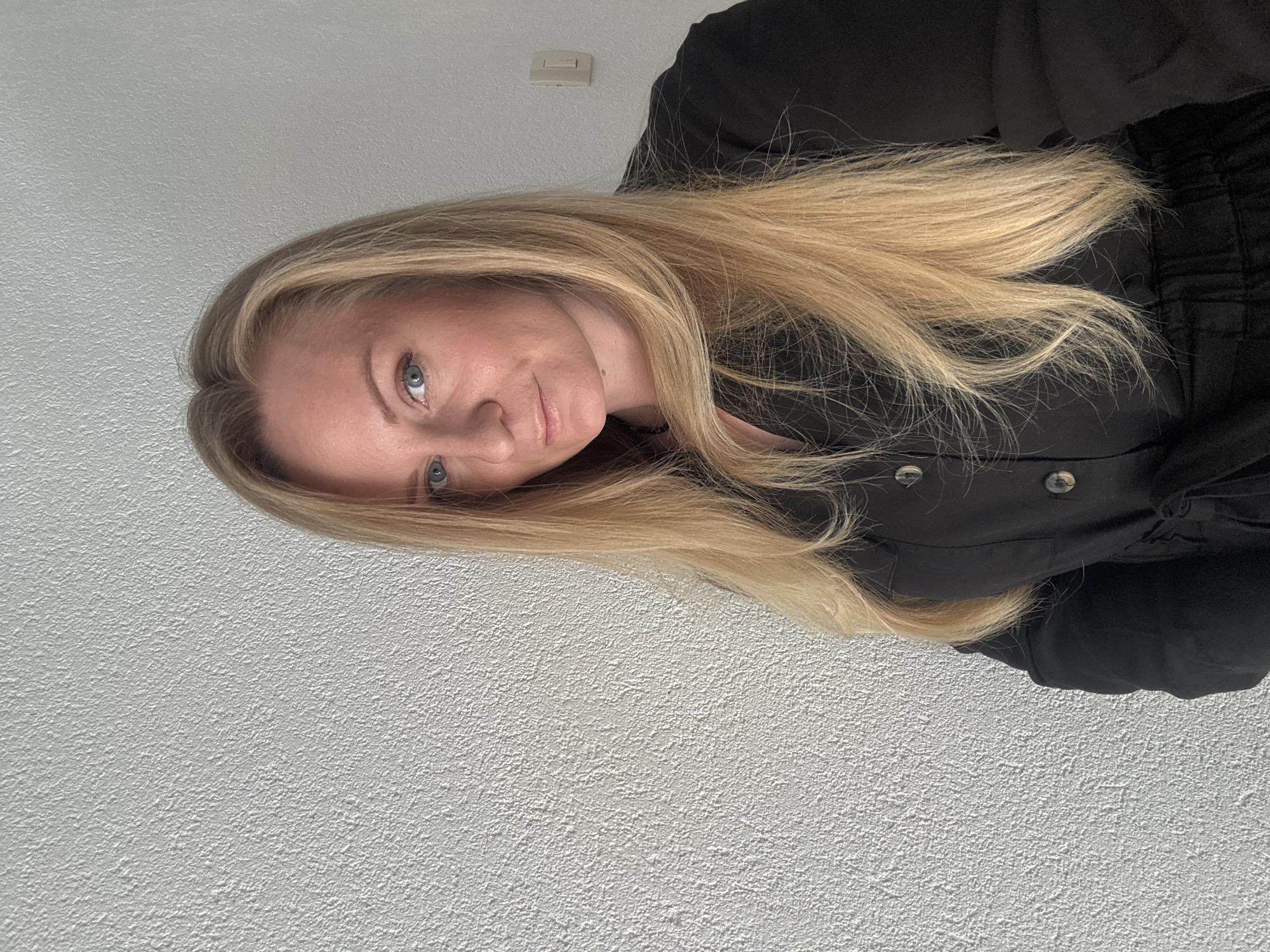 A woman with long, blonde hair and blue eyes poses in front of a white wall. She is wearing a black button-down shirt. The location appears to be indoors, possibly in a residential building. The city and specific location are unknown.