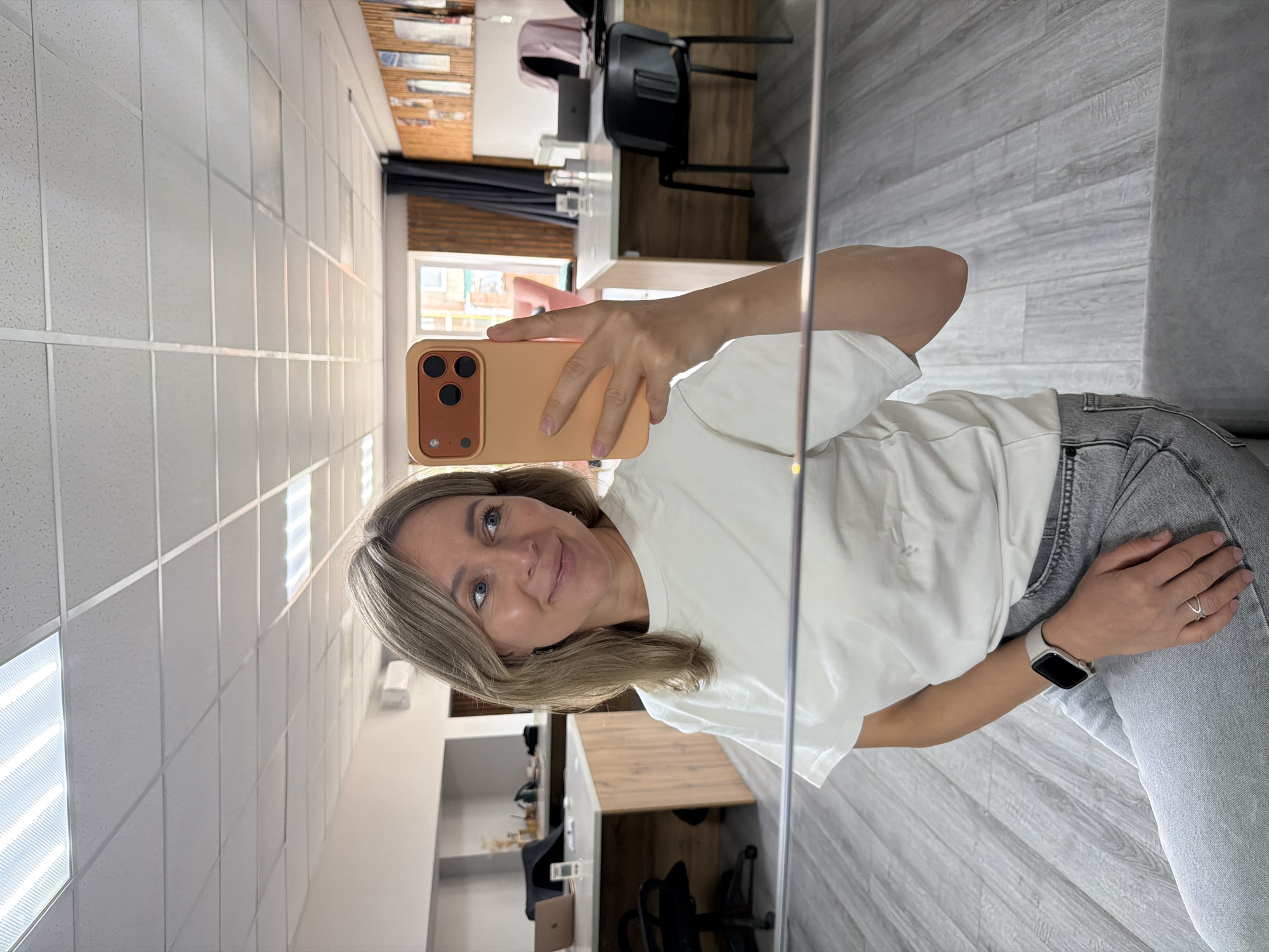 A woman is taking a selfie in an office. She is wearing a white t-shirt and grey jeans. She has blonde hair and is smiling. In her right hand, she is holding a peach-colored phone. In the background, there are desks, chairs, and a laptop. The floor is made of grey wooden planks. The ceiling has fluorescent lights. The overall impression is that of a modern office space. The woman is likely at her workplace in Chaikovsky, Russia.