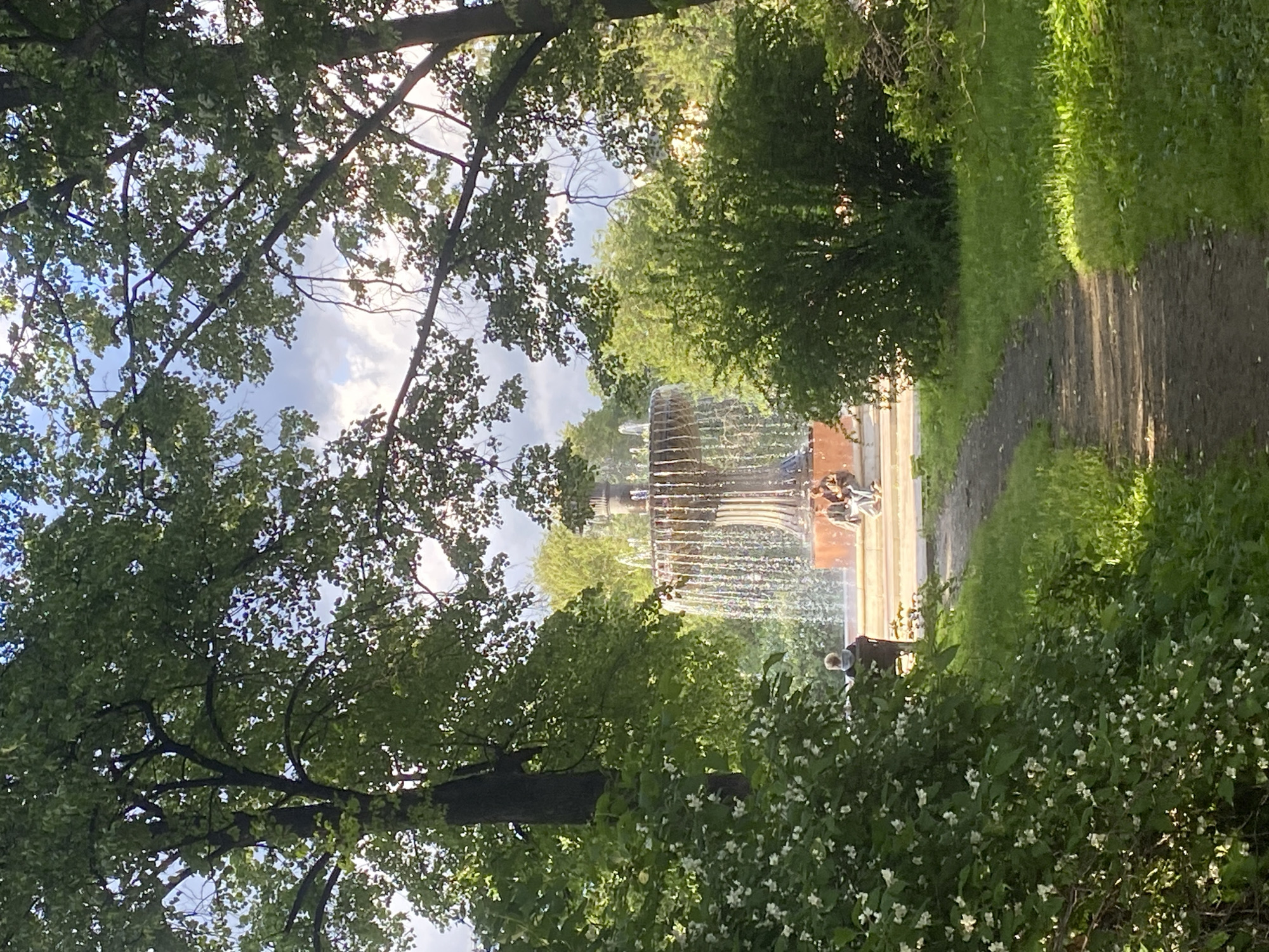 The image depicts a fountain in a park in Yekaterinburg, Russia. The fountain is multi-tiered with water cascading downwards. There are people sitting near the fountain and others walking on a path beside it. The scene is framed by trees and greenery, including some white flowering bushes in the foreground. The sky is visible through the trees.
