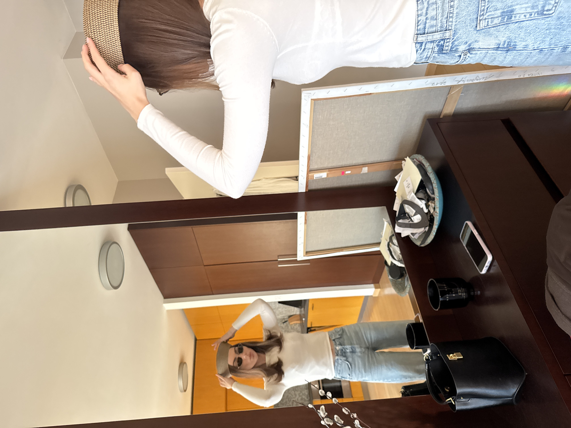 A woman wearing a white long-sleeved shirt and blue jeans is adjusting a beige hat on her head, as seen in a mirror reflection. The reflection shows a room with light-colored walls and circular wall-mounted light fixtures. The mirror is set into a dark wood frame, which also serves as a shelf or countertop. On the shelf, there is a black purse, a camera lens, a phone, and a bowl containing assorted items, including what appears to be shoes. An artwork stands on the shelf. The background of the reflection reveals a hallway with light brown wooden cabinets. The location is Unknown, Unknown.
