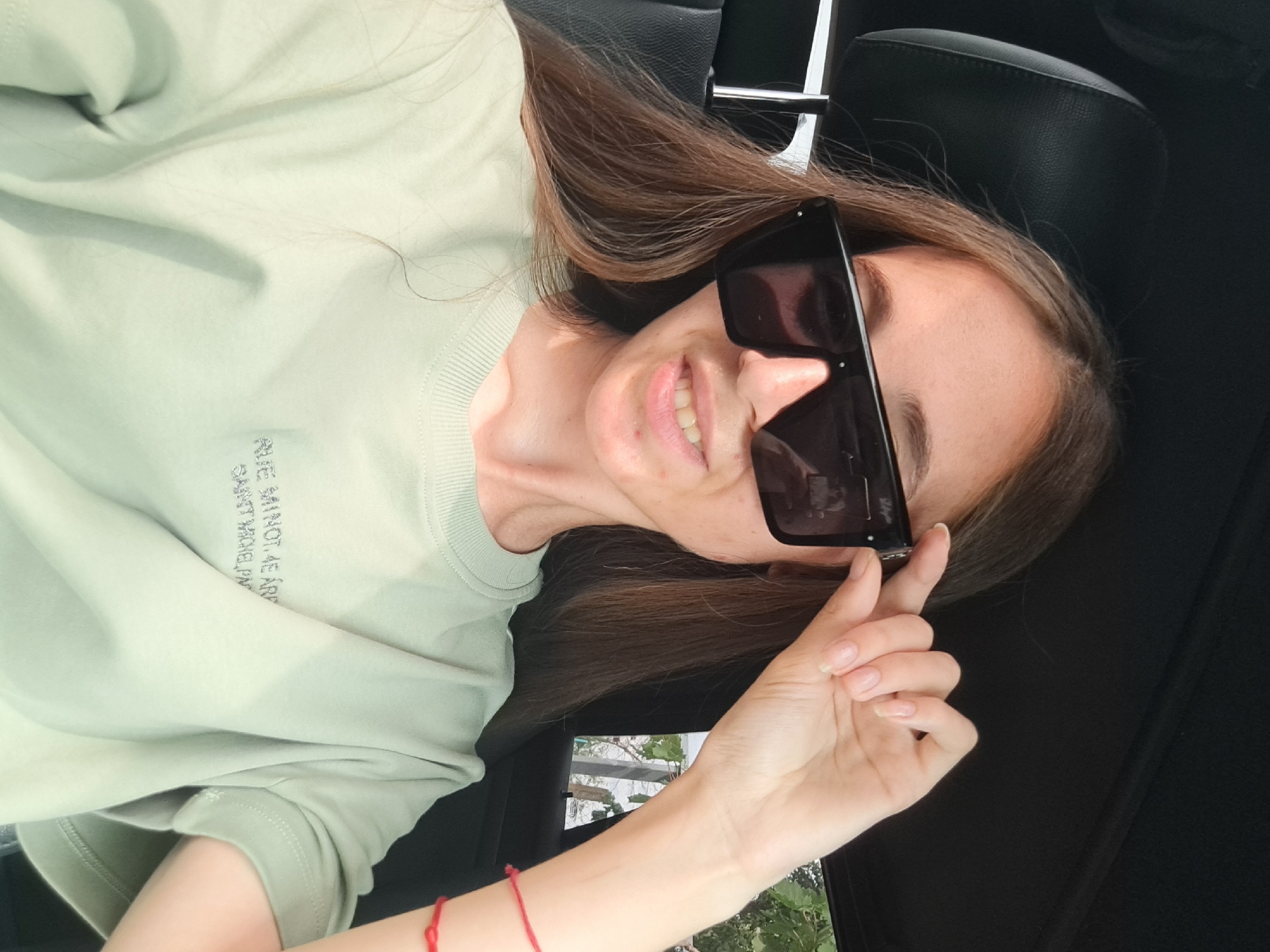 A young woman with long brown hair and wearing sunglasses and a light green t-shirt is sitting in a car. She is adjusting her sunglasses with her right hand. The t-shirt has a small, embroidered text in grey on the left side of the chest. A red string bracelet is visible on her right wrist. The background is out of focus but shows a glimpse of greenery and a fence. The scene appears to be outdoors, possibly in Vypasne, Ukraine, given the presence of a car and natural elements.