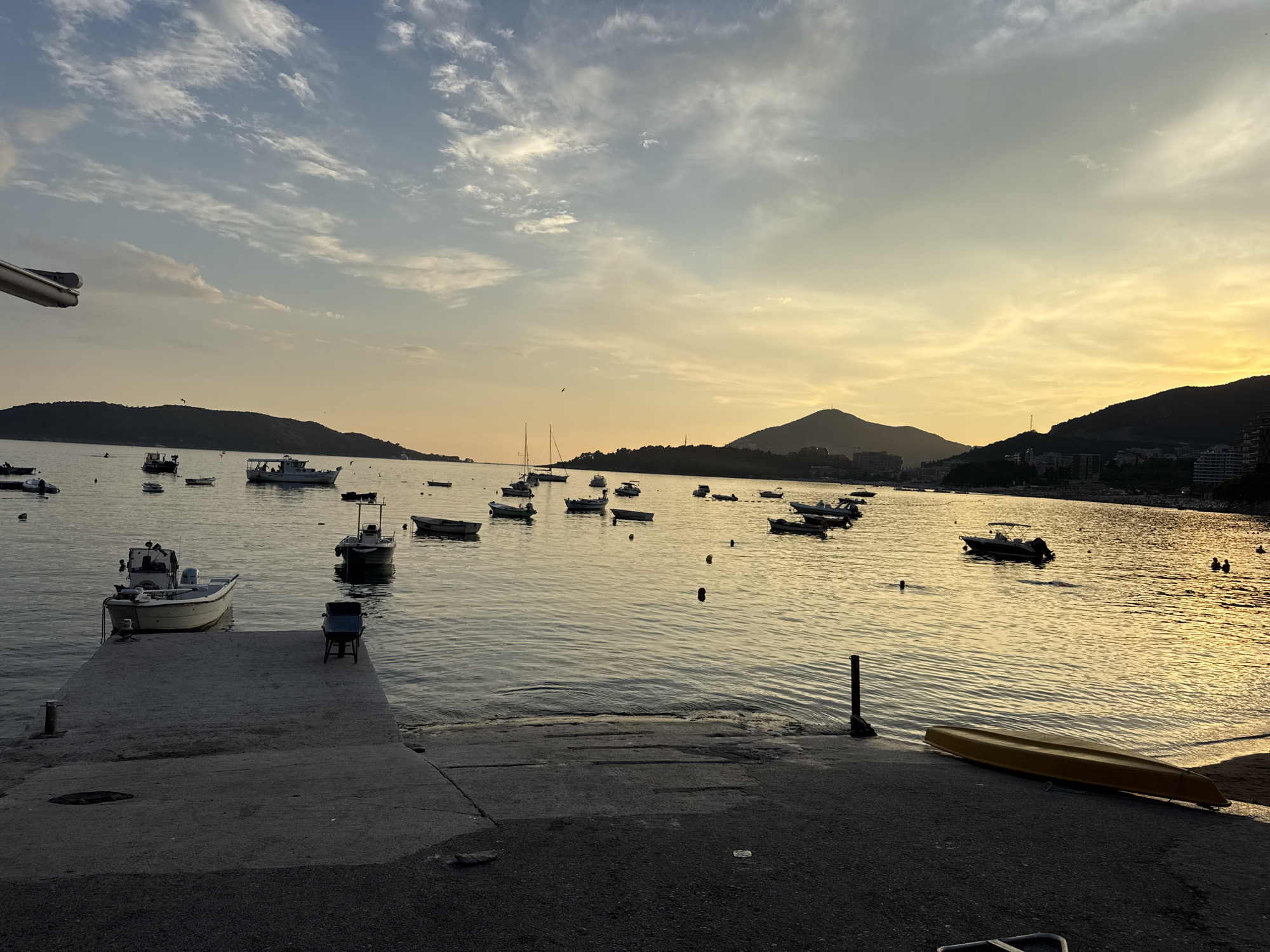 The image shows a serene sunset over the sea in Rafailovići, Montenegro. Several boats are anchored in the calm waters, some of which are small fishing vessels, while others appear to be leisure boats. The sky is a gradient of soft blues and yellows, with wispy clouds scattered across it, reflecting on the water's surface. In the background, the silhouette of the coastline is visible, with hills and some buildings suggesting a town or resort. In the foreground, a concrete pier extends into the water, with a yellow kayak lying on the shore beside it. The overall atmosphere is peaceful and tranquil, typical of a coastal evening.
