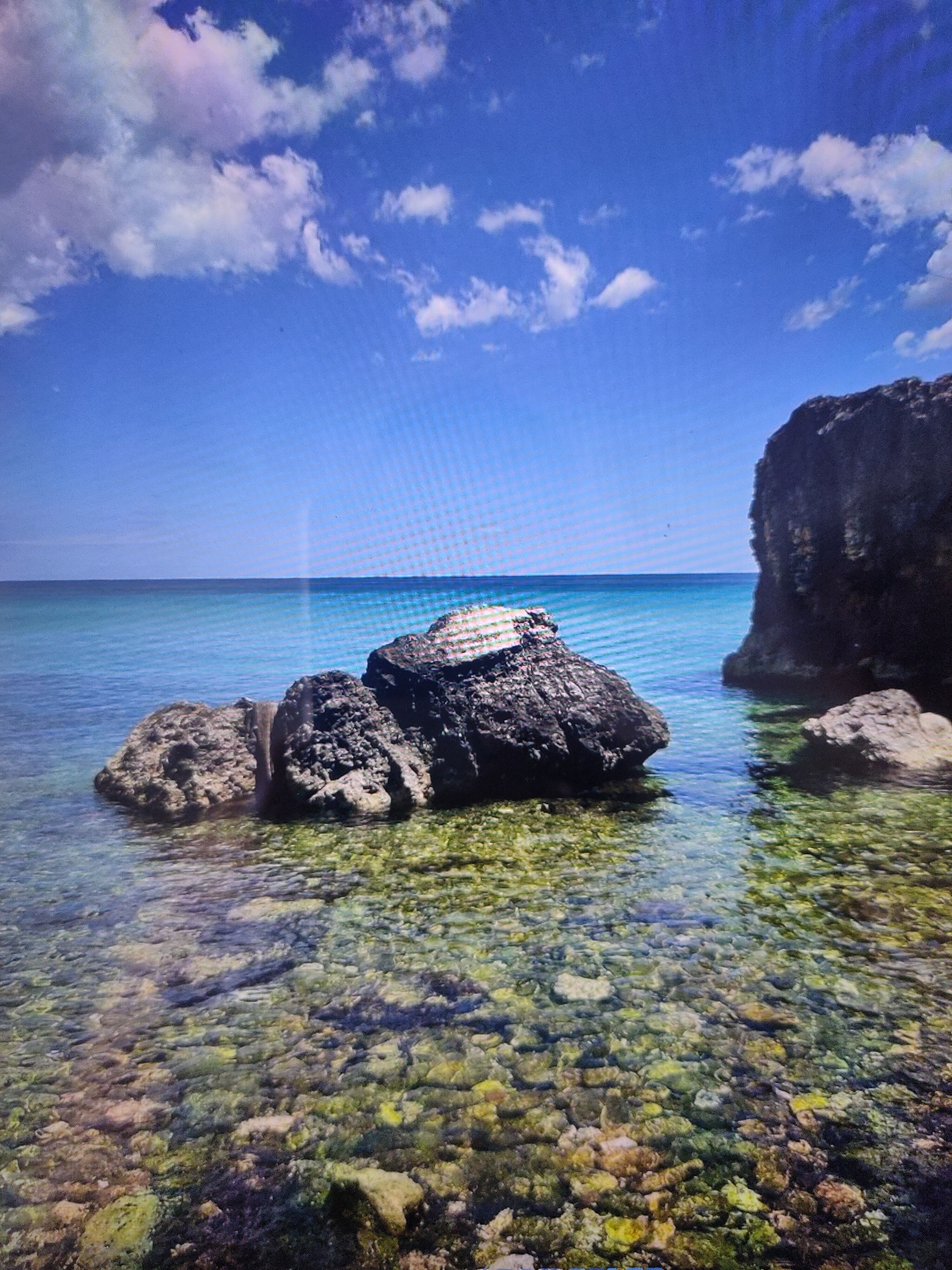 This is a photograph of a rocky coastline with clear turquoise water. In the foreground, several large rocks are partially submerged in the shallow water, revealing a seabed covered in pebbles and algae. Further out, the water deepens and becomes a vibrant blue under a clear sky dotted with white clouds. On the right side of the image, a large, dark rock formation juts out from the water. The scene depicts a natural, serene coastal environment. There are no people or animals visible in the image, and no indications of urban or man-made structures within the frame. The photograph does not appear to be taken in Zrenjanin, Serbia, as Zrenjanin is an inland city and does not have a coastline.