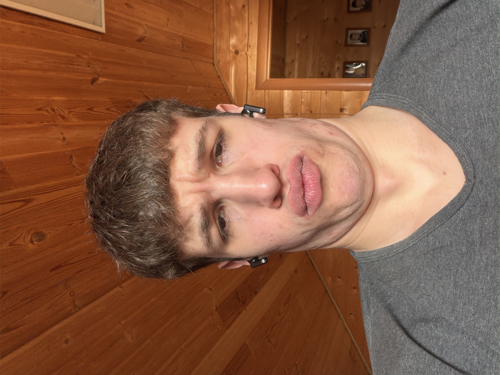 The image features a young man captured from the chest up, oriented sideways against a warm, wooden-paneled background. He has short, disheveled brown hair and wears a plain grey t-shirt. Black wireless earbuds are distinctly visible in both of his ears. The man is making a pronounced facial expression, characterized by furrowed brows, a wide-eyed gaze, and a downturned, puckered mouth, suggesting a grimace, pout, or an attempt at a humorous face. His skin exhibits some redness and minor blemishes on his cheeks and neck, with visible folds under his chin.
The background predominantly features rustic wooden paneling, indicative of an indoor setting like a sauna, a cabin, or a traditional wooden interior. In the upper right portion of the background, a mirror with a wooden frame reflects indistinct objects, possibly framed pictures, while a partial rectangular object (perhaps a light fixture or another frame) is visible in the upper left. A thin rope or cord runs vertically near the right side of the man's head. The overall setting implies an indoor environment, likely a home or facility in Прикамский, Russia. There is no visible text in the image.