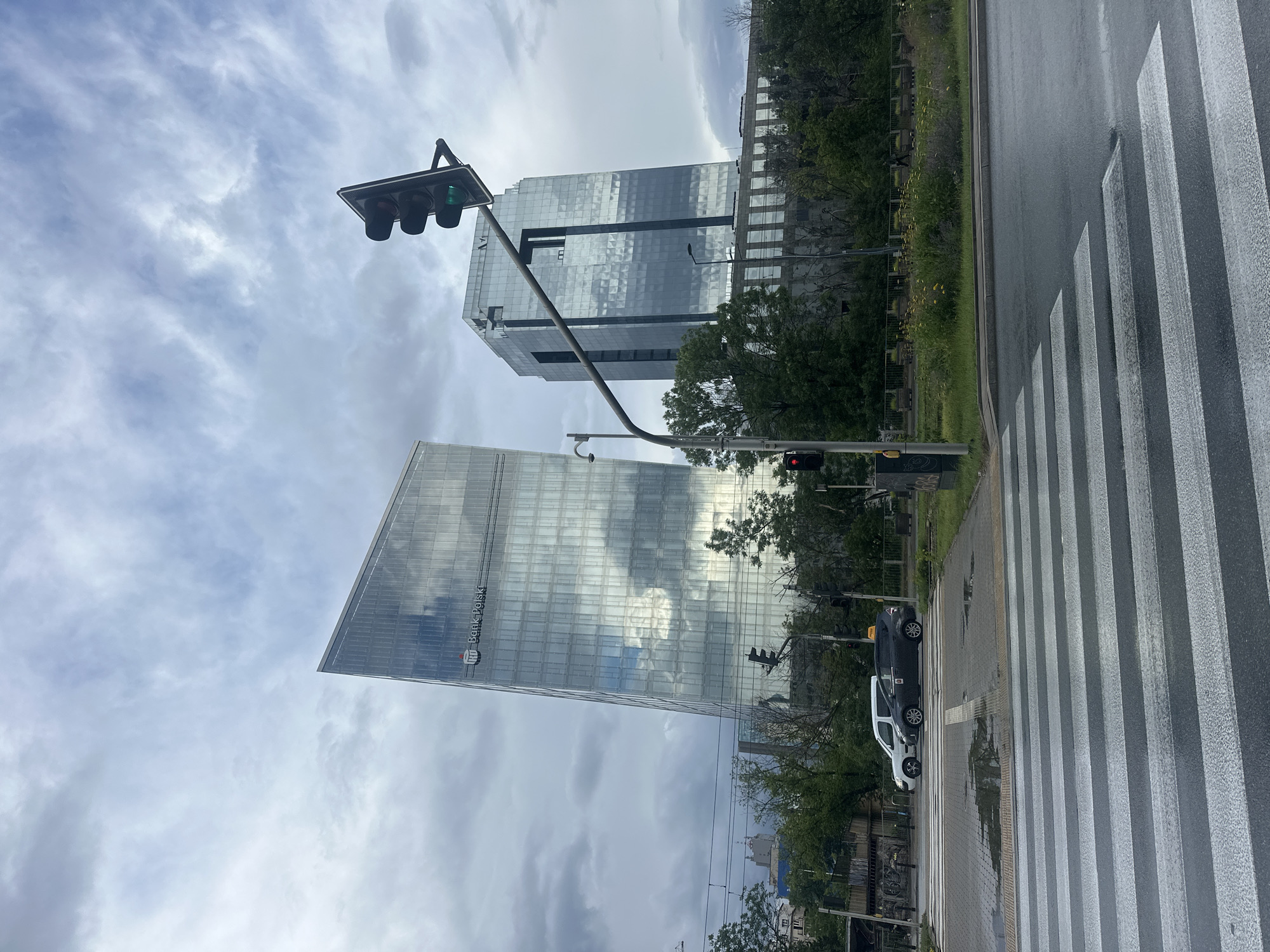 The image depicts a street scene in Warsaw, Poland, with a focus on two modern glass-faced skyscrapers and a pedestrian crossing in the foreground. The skyscraper on the left, visibly angled, displays the "Bank Polski" logo. The second skyscraper, to its right, is taller and more rectangular. A traffic light overhead shows a green light, with a red light visible on a separate pole further down the street. A pedestrian crossing with white stripes is wet from rain, with two cars, a white SUV and a dark minivan, stopped at the crossing. Trees and greenery line the background, with a cloudy sky overhead.