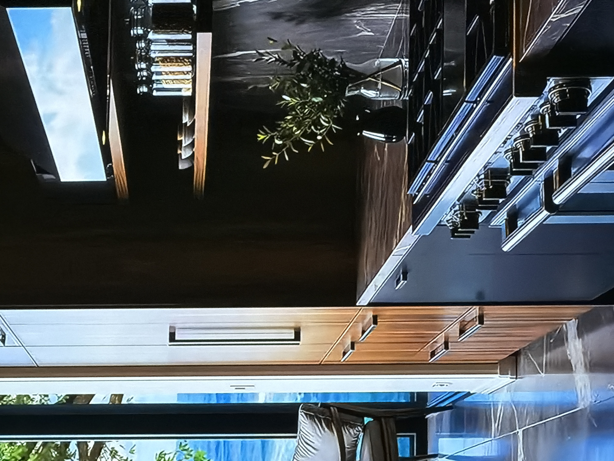 The image presents a high-end, modern kitchen interior, shown rotated 90 degrees counter-clockwise. When viewed upright, the scene reveals a sleek and sophisticated design.
A prominent feature is a large, professional-style range or stove with multiple burners and heavy-duty control knobs, positioned on the right side of the frame. Above it, a matching range hood is integrated into the cabinetry. The countertops and backsplash are crafted from a dark, polished stone, likely marble or granite, contributing to the luxurious feel. A vase with slender, green foliage is placed on the dark counter, adding a touch of natural elegance.
To the left of the cooking area, a multi-tiered open shelf or spice rack holds several glass jars with contents, suggesting organized storage. Above this, a long, narrow window or light fixture reflects a bright blue sky with clouds, indicating it is daytime. The kitchen features a combination of dark upper cabinetry and lighter-toned wooden lower cabinets, possibly oak or a similar wood, fitted with minimalist, horizontal handles.
Below the main counter, more wooden cabinets are visible, along with reflective surfaces that could be integrated appliances. At the bottom of the image, a large window or glass door looks out onto an outdoor scene with a clear blue sky and some greenery. A part of a chair or outdoor furniture is also visible through this window. The aesthetic is contemporary, characterized by clean lines, contrasting dark and light materials, and a sense of spaciousness. No specific activity is depicted, and there is no visible text. The setting is a well-appointed kitchen in what appears to be a residential property in Sofiivska Borshchahivka, Ukraine.