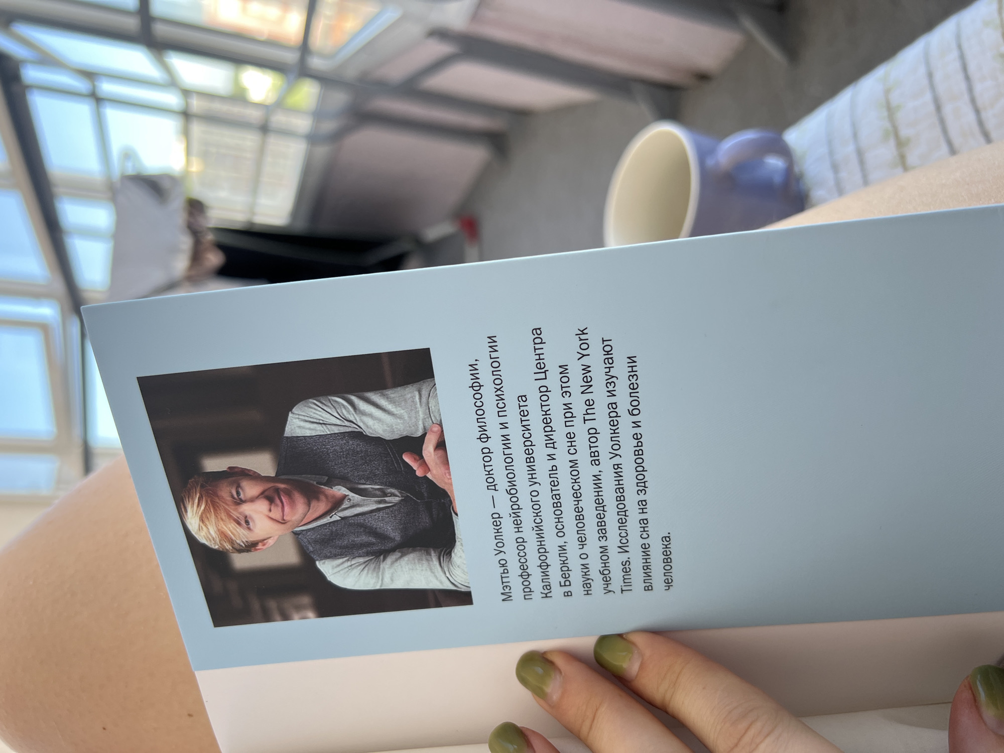 A person's arm and hand with green nail polish is in the foreground, holding open a light blue book. The book features a portrait of a blonde man in the upper left quadrant. Below the portrait, text is printed in Russian. The background shows a sunlit room with large windows, a purple mug, and a patterned cushion. The scene is indoors. There is no identifiable information about the location being Novosibirsk, Russia.