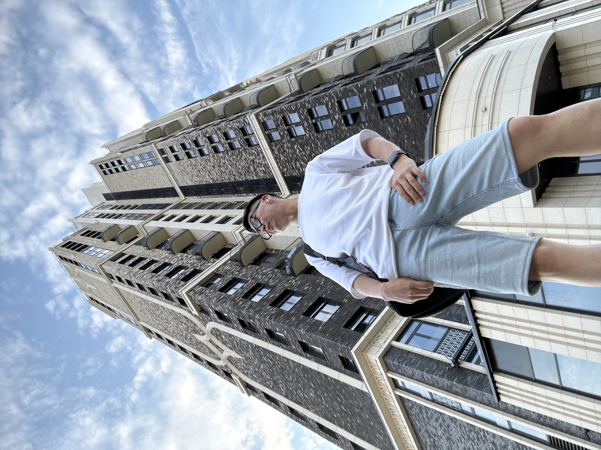 A person stands in front of a tall, modern building with a dark brick facade and light-colored accents. The building has many windows and balconies. The sky above is blue with scattered clouds. The person is wearing a white t-shirt, light blue denim shorts, glasses, and a watch. A black bag is slung over their shoulder. The perspective is from a low angle, looking up at the building and the person.