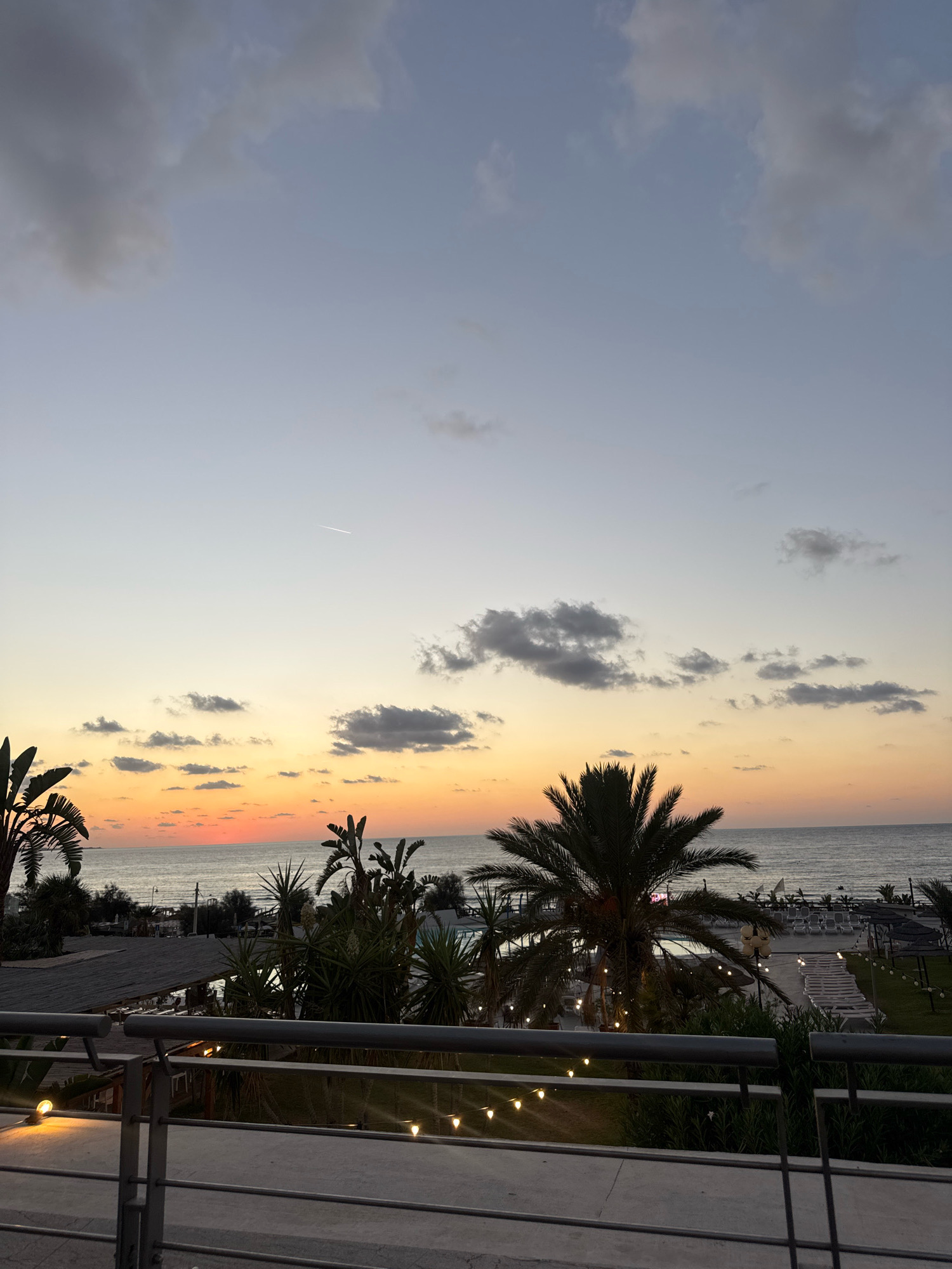 This is a photograph taken at sunset from an elevated viewpoint overlooking a resort or beachfront area. The sky displays a gradient of colors, transitioning from a pale blue at the top to shades of orange and pink near the horizon, indicating the setting sun. Several clouds are scattered across the sky, some appearing darker and more defined, while others are wispy. A faint contrail from an airplane is visible in the upper right portion of the sky.
In the middle ground, the sea is visible, with gentle waves. Along the coastline, there are palm trees and other tropical foliage, silhouetted against the sunset. Beyond the trees, there are glimpses of resort amenities, including lounge chairs, umbrellas, swimming pools, and buildings with thatched roofs. String lights are illuminated, casting a warm glow over the scene, particularly in the foreground and middle ground. A metal railing is prominent in the foreground, suggesting the viewpoint is from a balcony or terrace.
While the location is stated to be Isola delle Femmine, Italy, no specific landmarks are identifiable in the image to confirm this. There are no discernible people actively engaged in any actions within the frame. The overall atmosphere is serene and tranquil, characteristic of a sunset view at a vacation destination.
