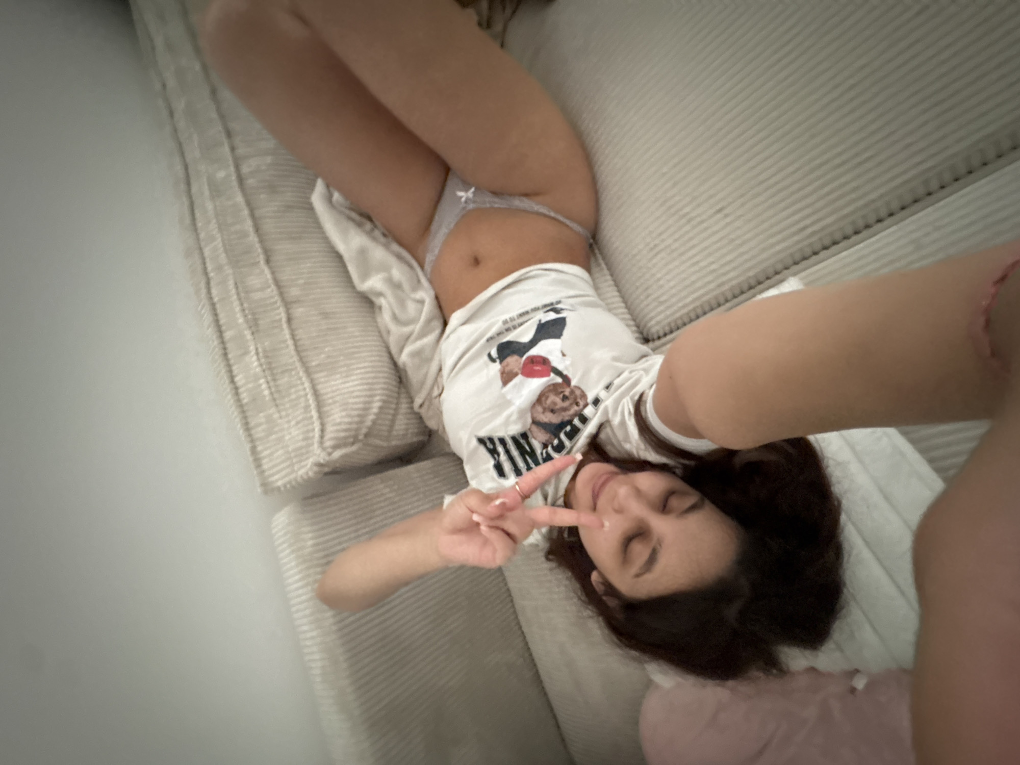 An overhead selfie captures a young woman lying diagonally on a light-colored, ribbed sofa or bed. She is wearing a white t-shirt featuring a cartoon graphic of a brown bear in a blue top and light grey underwear with a small white bow. Her midriff is visible.
Her eyes are closed or mostly closed, and she has a serene, relaxed expression. She holds her left hand up, making a peace sign, showcasing painted fingernails. Her dark hair is spread around her head, resting partially on a pink object, likely a pillow or blanket.
The background on the left is a soft, light grey wall, complementing the neutral tones of the sofa. The lighting is diffused and even, consistent with an indoor setting. Partial text is visible on her t-shirt, below the bear graphic, reading "YING". The overall impression is one of casual comfort and playfulness. No specific details within the image indicate the location as Mönchengladbach, Germany.