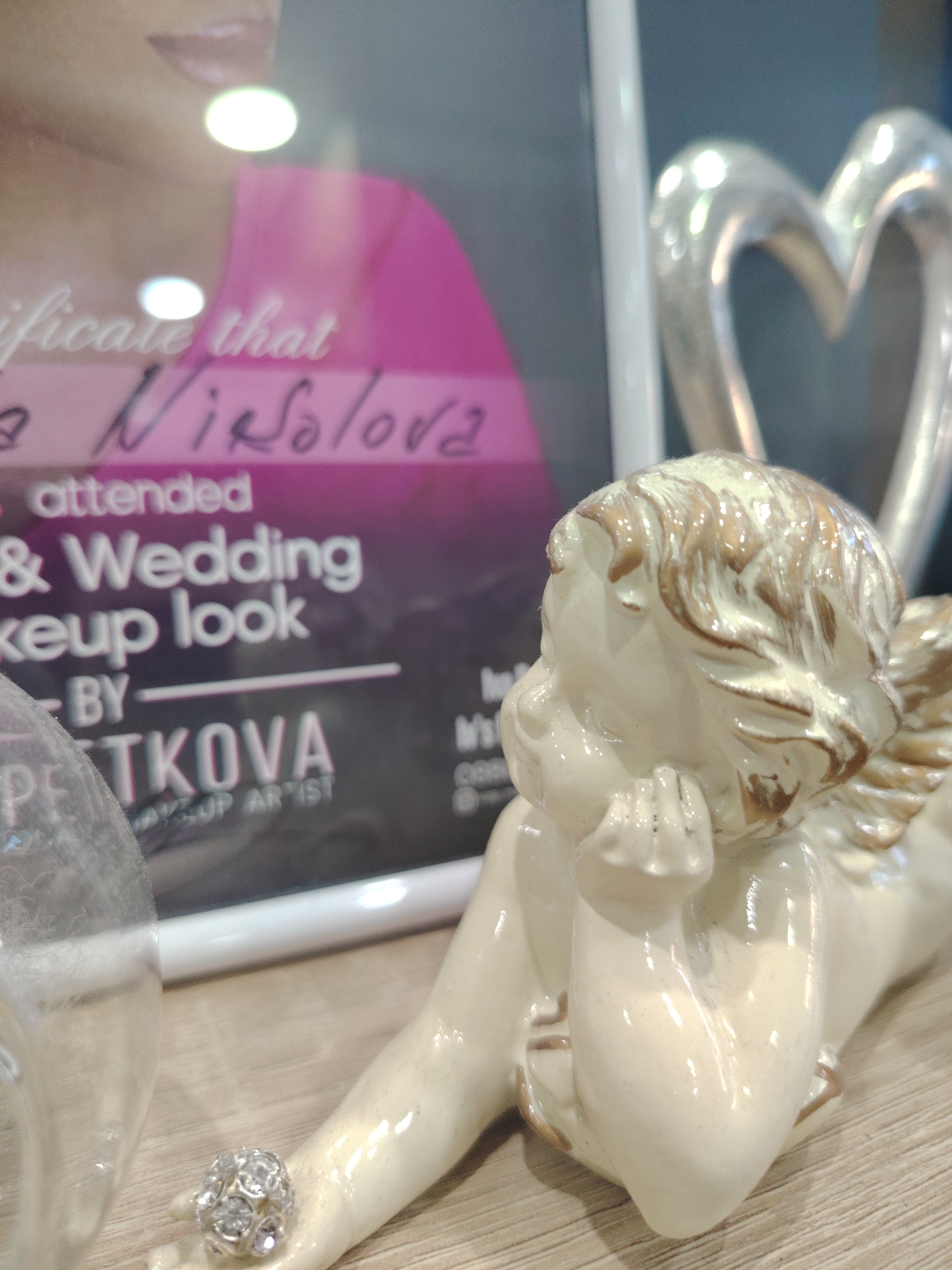 The image captures a close-up view of a small, decorative figurine of a winged cherub resting its head on its hand. The figurine is made of a pale, creamy material with gold accents highlighting its hair and wings. It is positioned on a light-colored wooden surface.
In the background, a framed certificate or document is partially visible, featuring text and an image of a woman's face and shoulder, with a bright pink garment. The text on the certificate appears to include phrases like "certificate that" and names. Also in the background, to the right, is a metallic sculpture of a heart.
To the left of the cherub figurine, a clear, rounded object, possibly a glass or plastic container, is present, obscuring part of the background document. A small, circular, gem-encrusted object, resembling a ring or embellishment, is placed on the cherub's outstretched hand.
This scene is located at the Makeup Artist Studio of Desislava PETKOVA, located in Haskovo, Bulgaria. The studio is situated at 78 Ivan Vazov Street, Haskovo.
