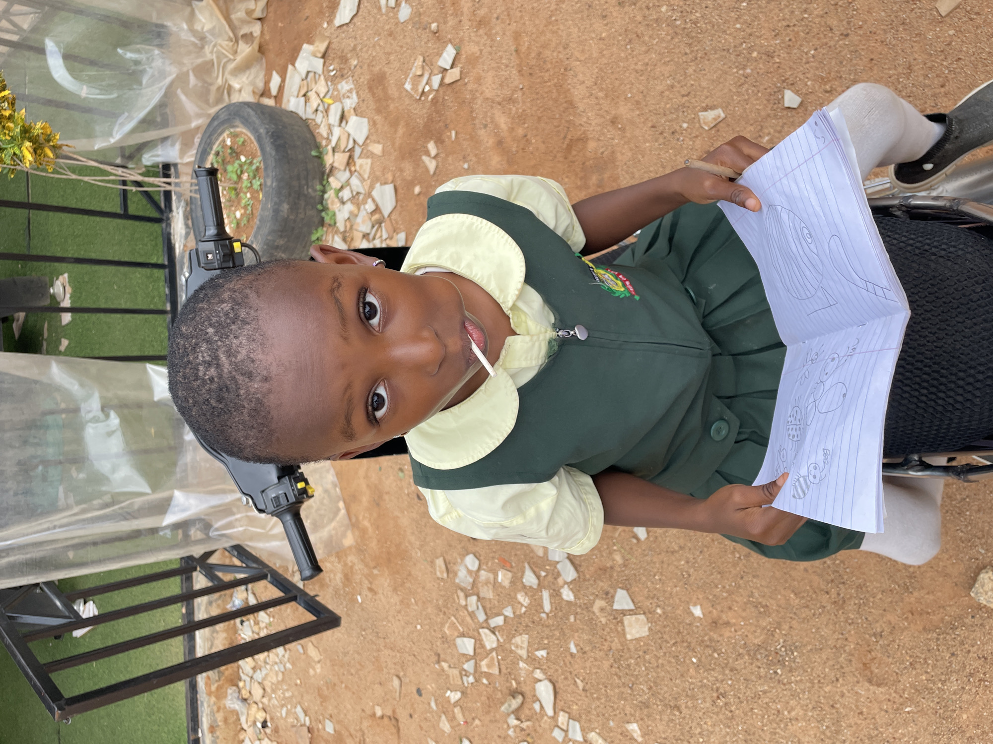 A young girl, likely of elementary school age, sits on the seat of a parked, small motorcycle, possibly a moped. She wears a school uniform with a dark green dress and light yellow collar and sleeves. In her hands, she holds a piece of lined paper with cartoonish drawings on it. She is looking up towards the camera, with a lollipop in her mouth.
The motorcycle is parked outdoors on a dirt surface interspersed with fragments of broken white tiles. Nearby are a rusty metal frame and an old car tire repurposed as a planter.
While the prompt specifies the location as Jos, Nigeria, there is no clear, definitive evidence visible in the image itself to confirm this, although the environment is consistent with images of semi-urban or rural environments in Nigeria.