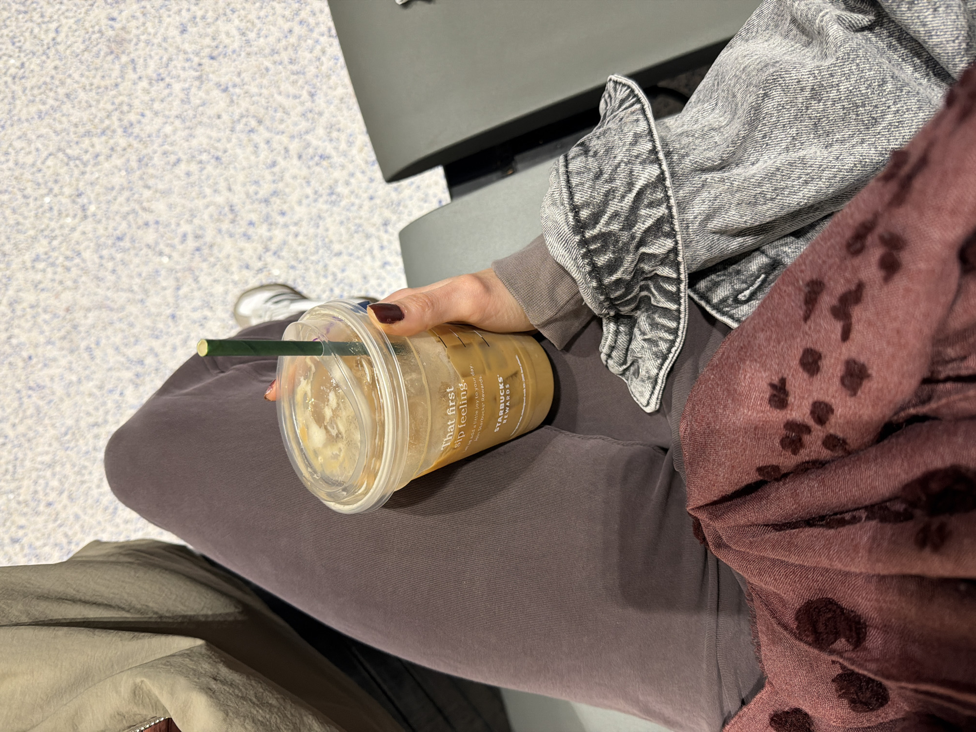 Here is a detailed description of the image:
The image shows a person sitting in what appears to be an airport waiting area. The person is holding a Starbucks iced coffee drink in their left hand. The cup has a green straw and is labeled with Starbucks branding and the phrase "That first sip feeling."
The person is wearing a gray acid-wash denim jacket, a brown sweatshirt underneath, and brown pants. A burgundy scarf with a dark brown pattern is draped on their lap. Part of what looks like another jacket is visible on the left bottom corner of the image.
The background is a gray speckled floor and a gray bench or seat. A shoe is visible on the left corner.