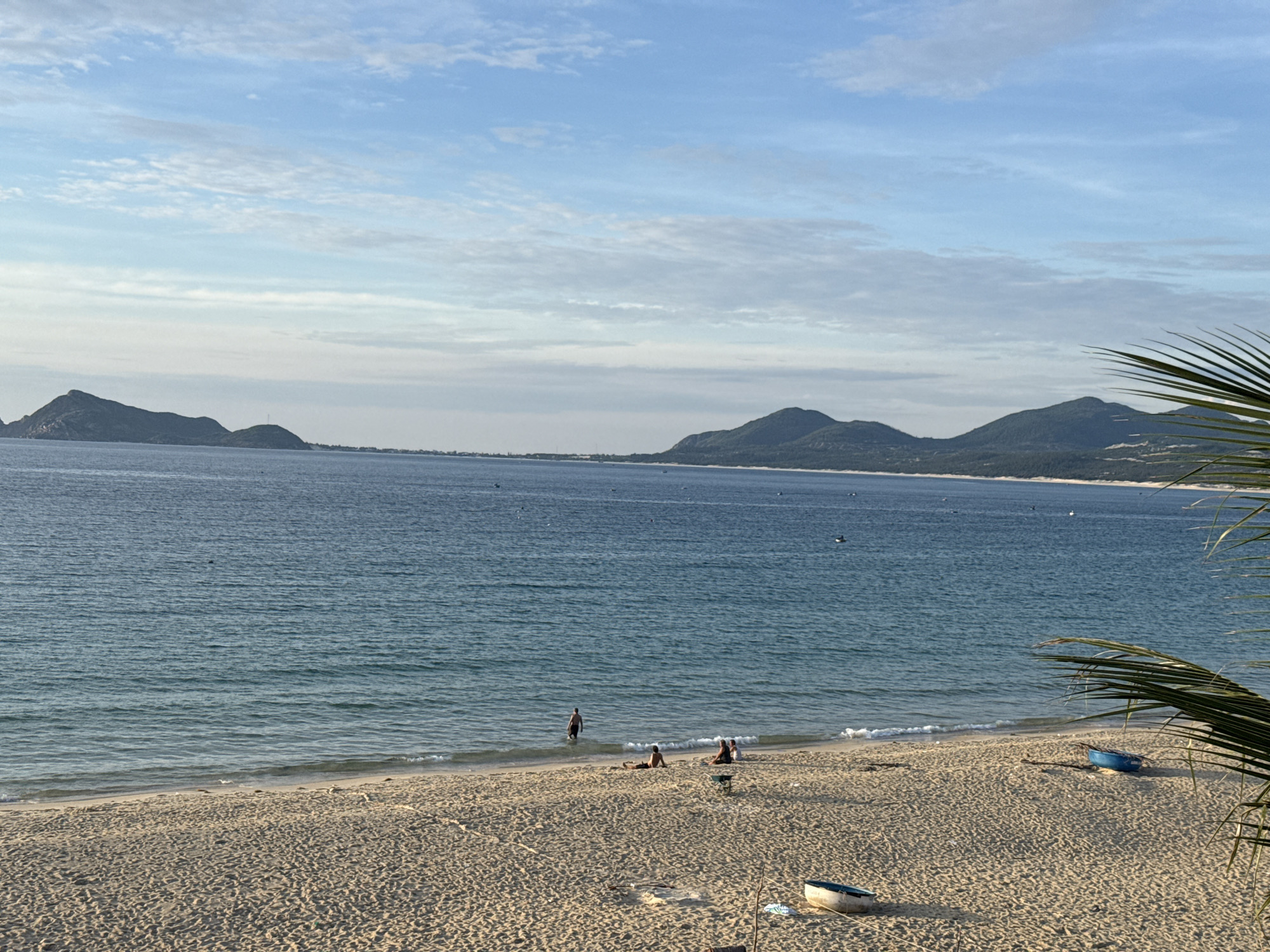 This image captures a scenic view of a tropical beach in Song Cau, Vietnam. The foreground is dominated by a sandy beach with scattered debris and two small boats, one blue and one white, resting on the shore. In the middle ground, the calm, blue ocean meets the shoreline, with gentle waves lapping at the sand. Several people are visible on the beach and in the shallow water. One person stands in the water, while others are seated or lying on the sand. The background features a distant coastline with lush, green hills and mountains under a bright blue sky dotted with white clouds. A palm frond partially frames the right side of the image. The overall atmosphere is one of tranquility and a leisurely day at the beach.