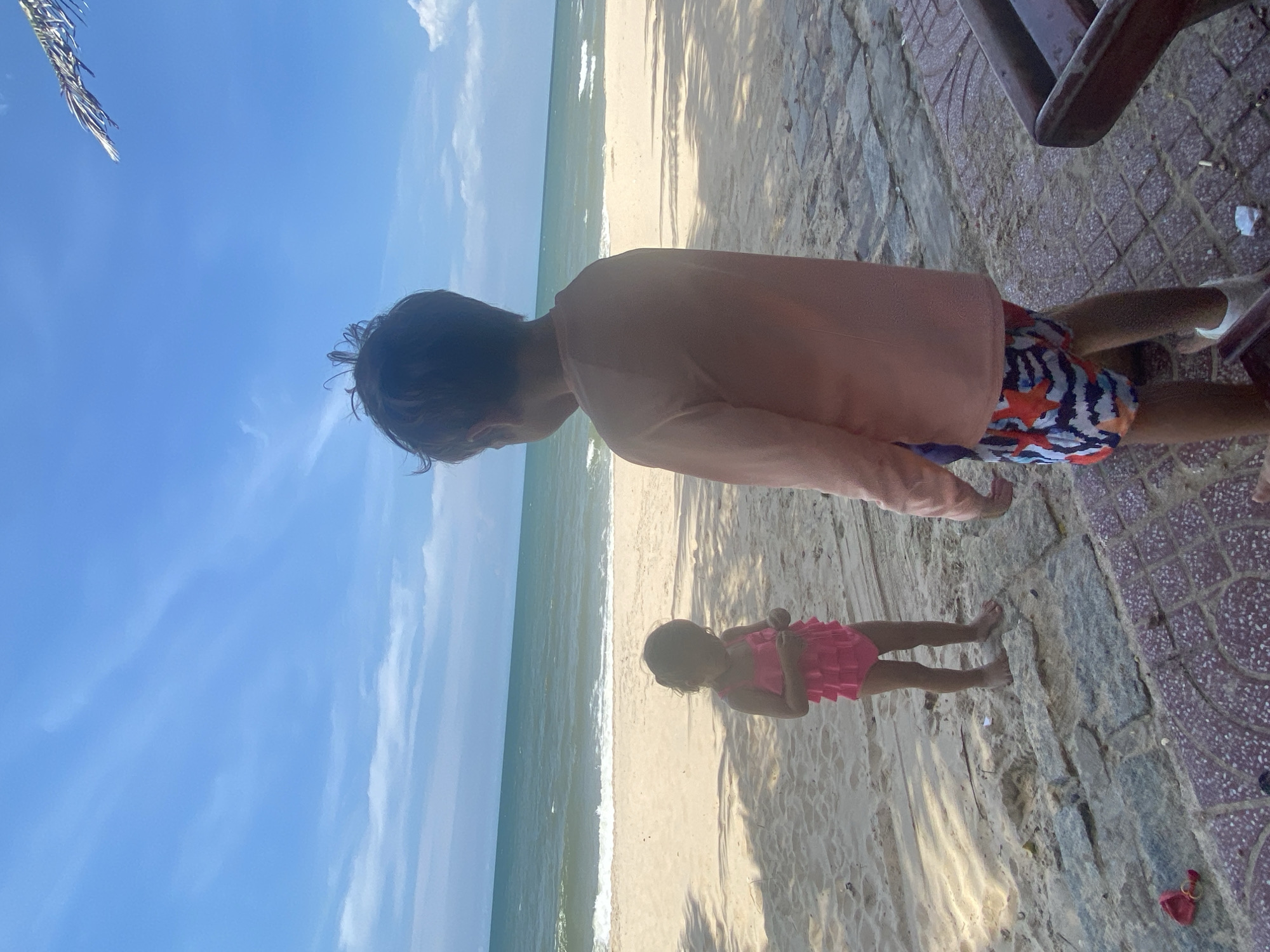 The image depicts a beach scene with two individuals. A person, likely an adult, is wearing a long-sleeved pink shirt and patterned swim trunks. Their back is to the camera, and they are standing on a sandy area next to a tiled path. To their right, partially visible, are the legs of a chair or bench. In the foreground, a young child, dressed in a pink ruffled swimsuit, is standing on the sand. The child's back is also to the camera, and they appear to be looking towards the ocean.
The scene is set on a beach with the ocean visible in the background, characterized by gentle waves and a sandy shoreline. The sky above is a clear blue with some scattered clouds, suggesting a sunny day. Shadows cast on the sand indicate the sun is present, likely mid-day. The tiled path suggests a developed area adjacent to the natural beach, possibly a promenade or walkway. The overall mood appears relaxed and leisurely, fitting for a beach outing.
The location is identified as Phu Thuy Ward, Vietnam. There is no visible text in the image.