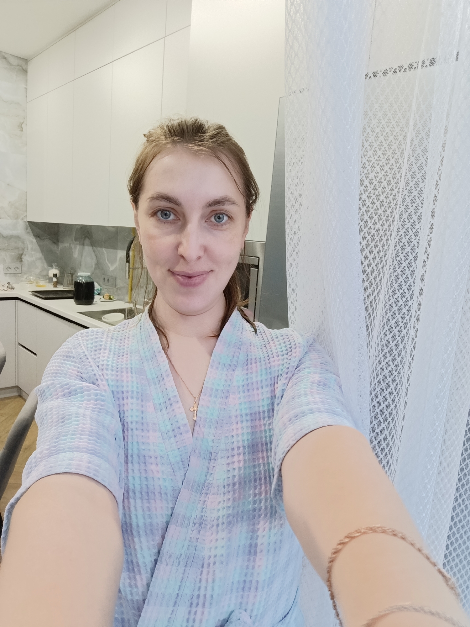 A medium shot depicts a fair-skinned woman in a light-colored, patterned robe. She has blue eyes and her hair is pulled back from her face. A gold cross necklace is visible at her throat. She appears to be taking a selfie.
Behind her, the scene transitions to an interior. To the left, modern white kitchen cabinets are visible, suggesting a kitchen area. A stainless steel sink and countertop with various kitchen items are partially visible. To the right, a white patterned curtain hangs.
The city of Chaikovsky, Russia is not evident in the image.