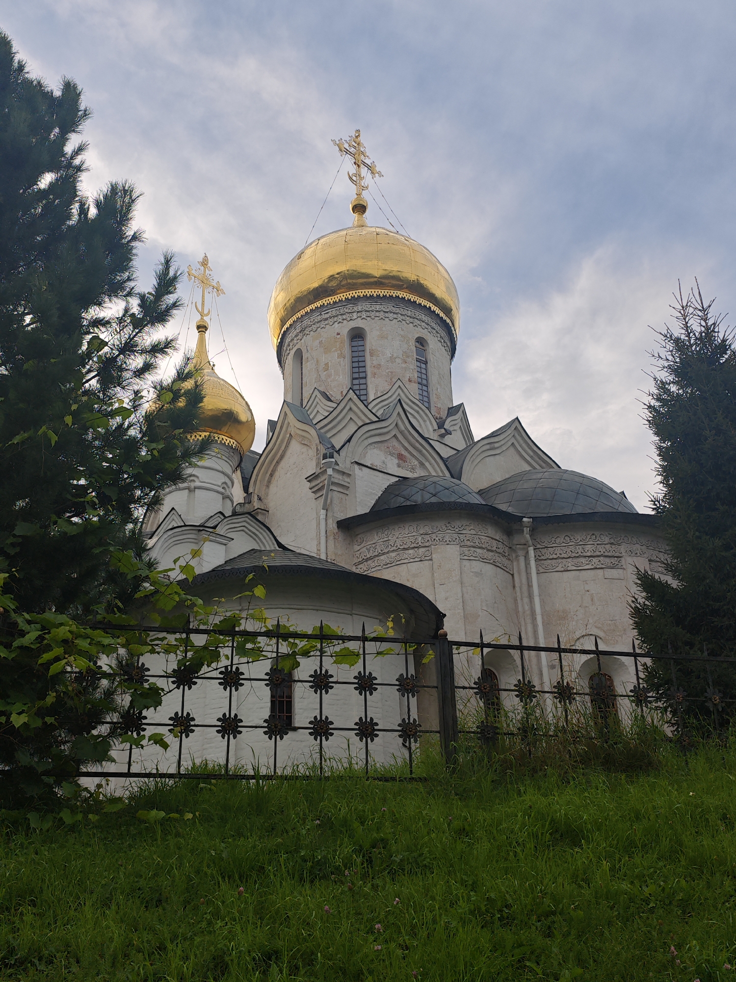 The image shows a low-angle view of the Ascension Cathedral in Zvenigorod, Russia. The cathedral, a white stone building with golden domes and crosses, is partially obscured by trees and a black metal fence in the foreground. Green grass covers the sloping ground in front of the fence, with a few small pink flowers visible. The sky above is cloudy, with patches of blue. The architecture of the cathedral is a classic example of pre-Mongol Russian church building, with prominent onion domes and intricate detailing. The scene appears peaceful and serene, typical of a religious site.
