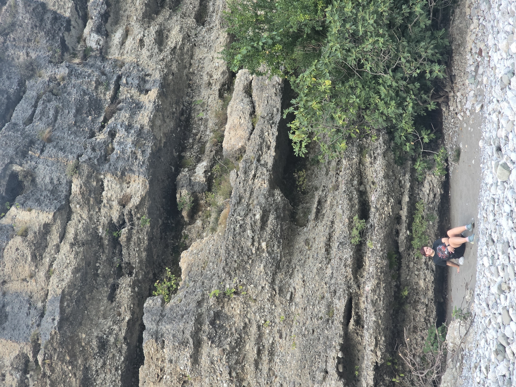 In the city of Bioče, Montenegro, a young person with short brown hair and a black t-shirt sits on a sandy patch at the base of a large, stratified rock formation. The rock face displays distinct horizontal layers of grey and beige material, with visible pebbles and some sparse vegetation growing from crevices. To the right of the rock formation, a dense green bush with numerous leaves and some small fruits is prominent. The foreground is covered with smooth, grey and white river stones. The person is facing towards the camera, with their legs extended and crossed, and their right arm resting on their knee. They are wearing dark shorts and teal-colored shoes. The overall scene depicts a person relaxing in a natural environment next to a rocky cliff face.