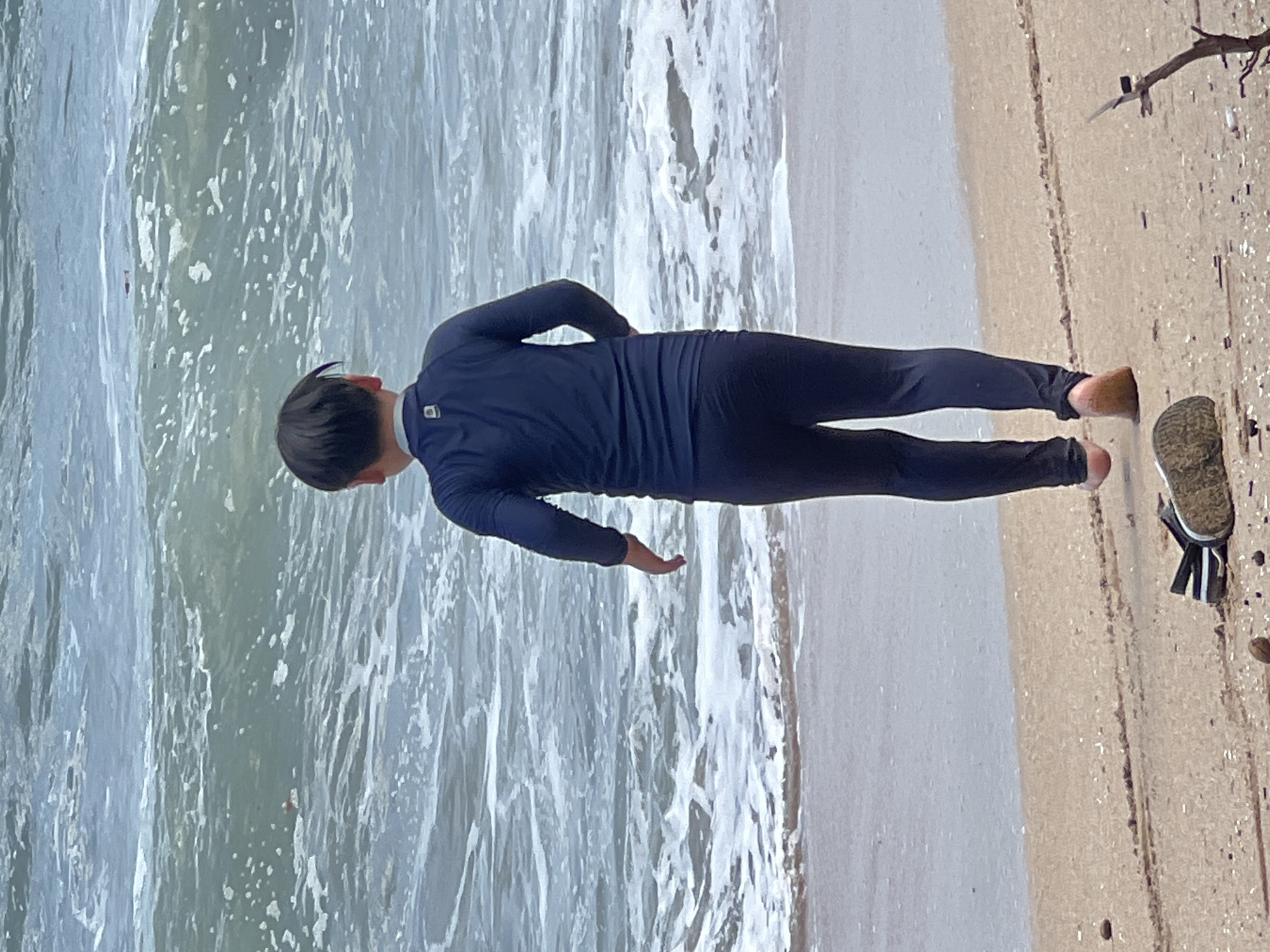A young boy wearing a navy blue rash guard and dark leggings stands on a sandy beach facing the ocean. The water is choppy with white foam and small waves rolling onto the shore. A pair of flip-flops lies on the sand in the foreground. The scene is overcast, suggesting a cloudy day. The location is the city of Kuta, Indonesia.