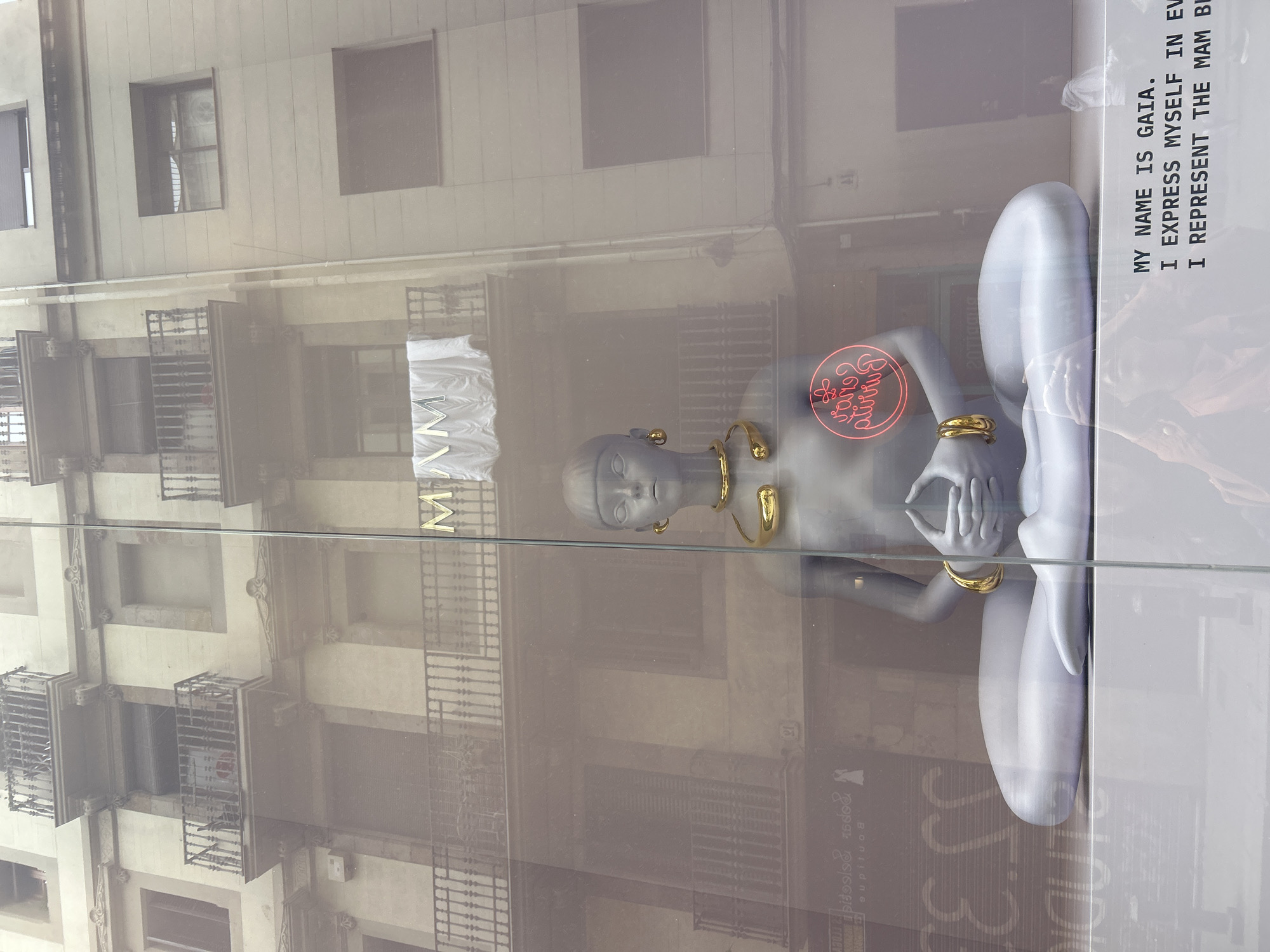 The image shows a display window in Barcelona, Spain, likely a storefront. The display features a pale gray mannequin in a seated, meditative posture, adorned with gold jewelry (earrings, a necklace, and bracelets). A red neon sign with reversed text is positioned near the mannequin's torso. To the right, text is printed on the window: "MY NAME IS GAIA. I EXPRESS MYSELF IN EV I REPRESENT THE MAM BI". The window reflects the buildings across the street, including windows and wrought-iron balconies. A sign with text, reversed from the viewpoint, appears at the bottom of the glass.
