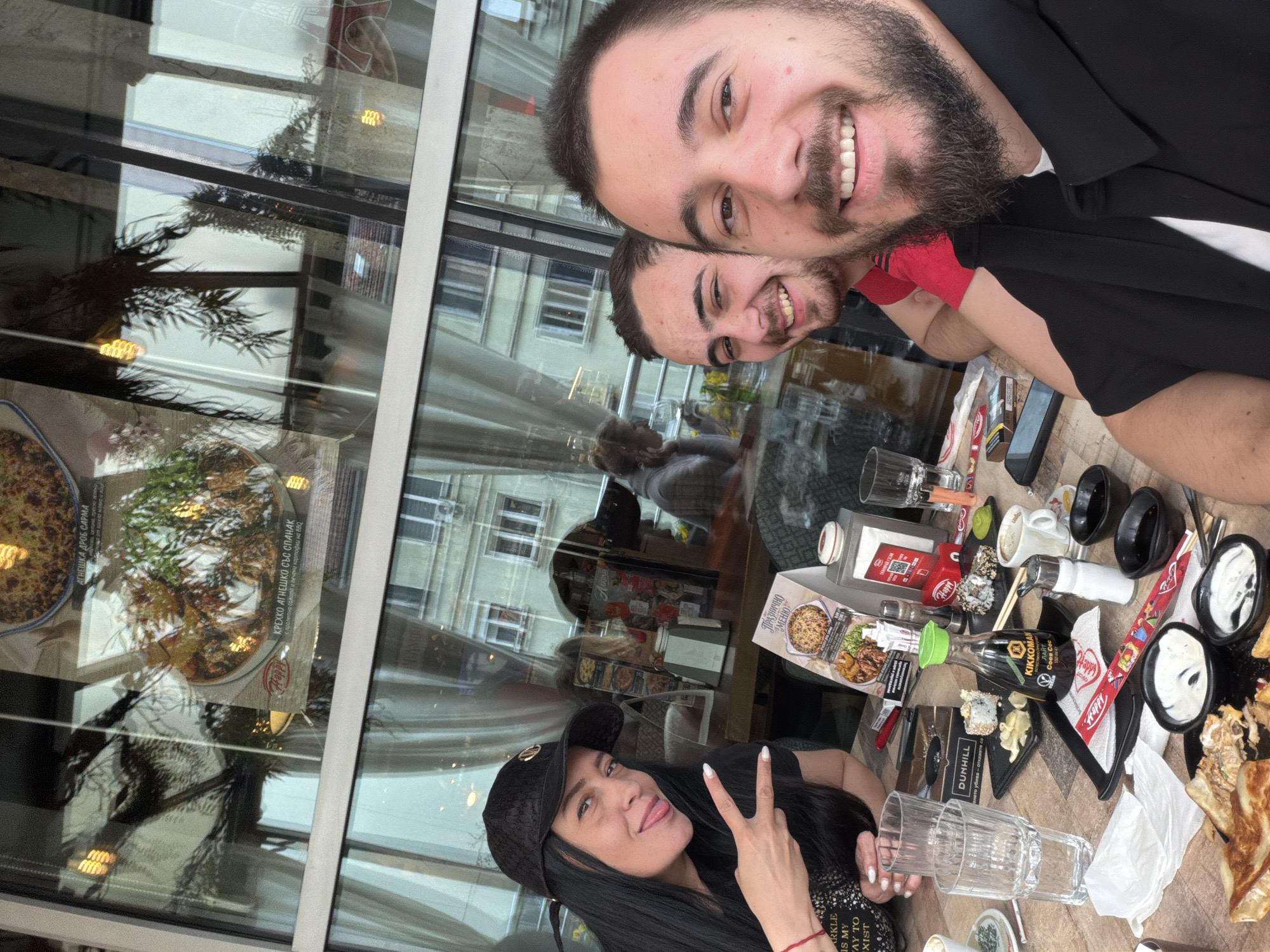 Here is a detailed description of the image:
**Scene:**
The image captures a group of three people dining at an indoor restaurant, likely in Sofia, Bulgaria. The photograph is taken from a slightly overhead, selfie-style perspective, featuring two men in the foreground and a woman seated across the table.
**People:**
* **Man 1 (Foreground):** He is smiling broadly at the camera, showing his teeth. He has dark hair, a beard, and is wearing a black shirt.
* **Man 2 (Behind Man 1):** He is partially visible, also smiling.
* **Woman:** She is seated, facing the camera and sticking out her tongue while making a peace sign with her fingers. She is wearing a black cap and a black top.
**Objects on the Table:**
The table is filled with food items and various containers, suggesting a shared meal. Notable items include:
* Bottles of Kikkoman soy sauce and other condiments.
* Small black bowls with white sauce
* Clear glass cups
* A box, possibly with menu information
**Environment:**
The scene is set within an indoor restaurant near a large window. Outside, the reflection of a building suggests an urban setting.
**Additional Notes:**
* The presence of specific condiments and labels (e.g., Kikkoman, "Happy") suggests possible information about the restaurant type or cuisine.
* Text from the menu reads "AгНЕШКА ДРОБ CАРМА" and "КРЕХКО АГНЕШКО СЪС СПАНАК" which translates to "Lamb liver sarma" and "Tender lamb with spinach".
* The composition and casual nature of the image suggest a candid moment shared among friends or family.